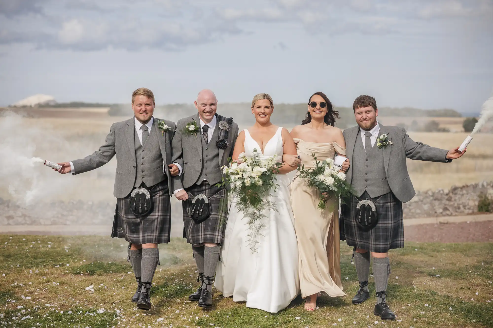 Four people in formal attire walk outdoors on grass, smiling: two men in grey jackets and kilts hold white smoke sticks, a bride in a white gown, and a bridesmaid in a beige dress. They carry floral bouquets. Rural landscape and blue sky appear in the background.