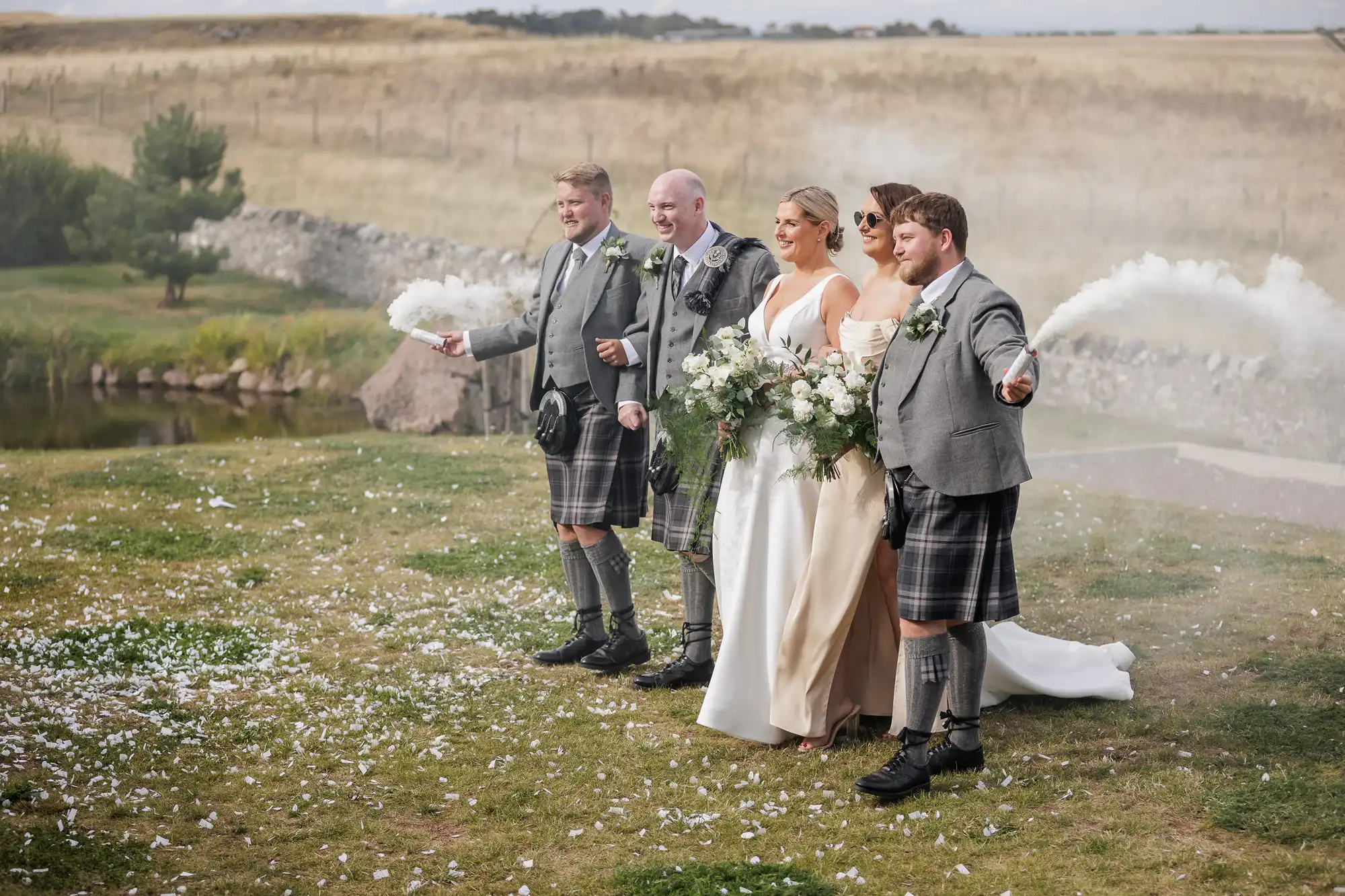A bride in a white dress and three men in grey jackets and tartan kilts stand outdoors on grass, posing for a photo. The bride holds a bouquet. The men spray white smoke from canisters. Flower petals cover the ground; fields and a pond are in the background.