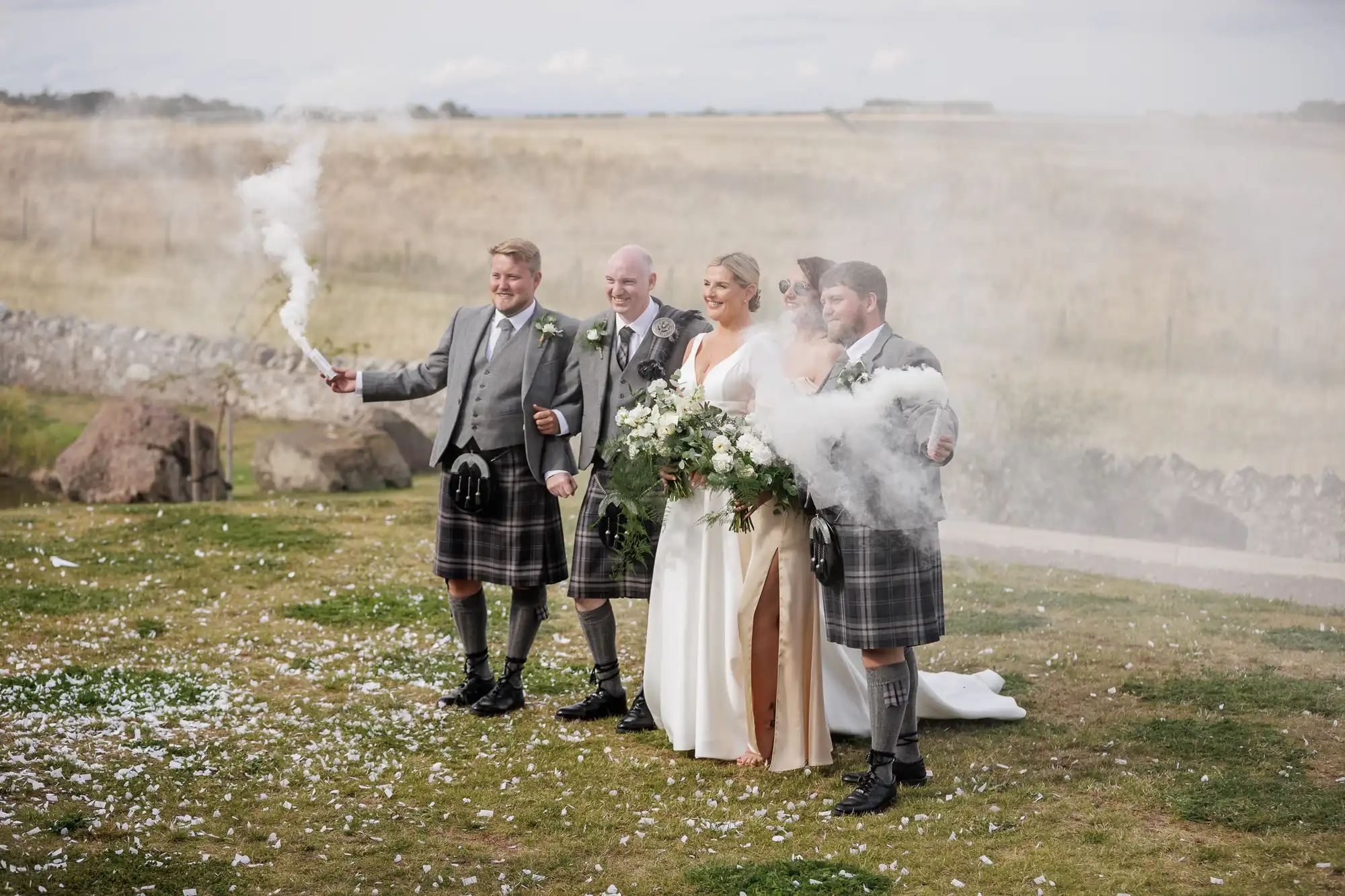 Five people in wedding attire stand outdoors on grass, smiling. Three men in grey kilts and jackets flank two women in white dresses holding large bouquets. Two men hold smoke bombs, with white smoke drifting around them. There’s a countryside landscape in the background.