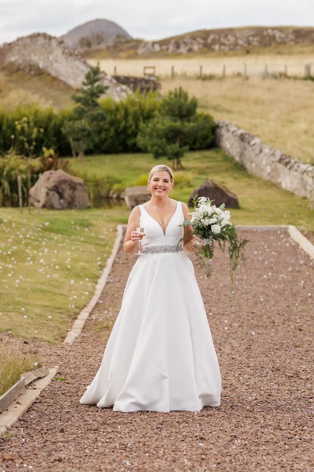 A smiling bride in a white gown and jewelled belt stands on a gravel path outdoors, holding a bouquet of white flowers and a glass. Behind her are green hills, a stone wall, bushes, rocks, and a distant mountain under a cloudy sky.