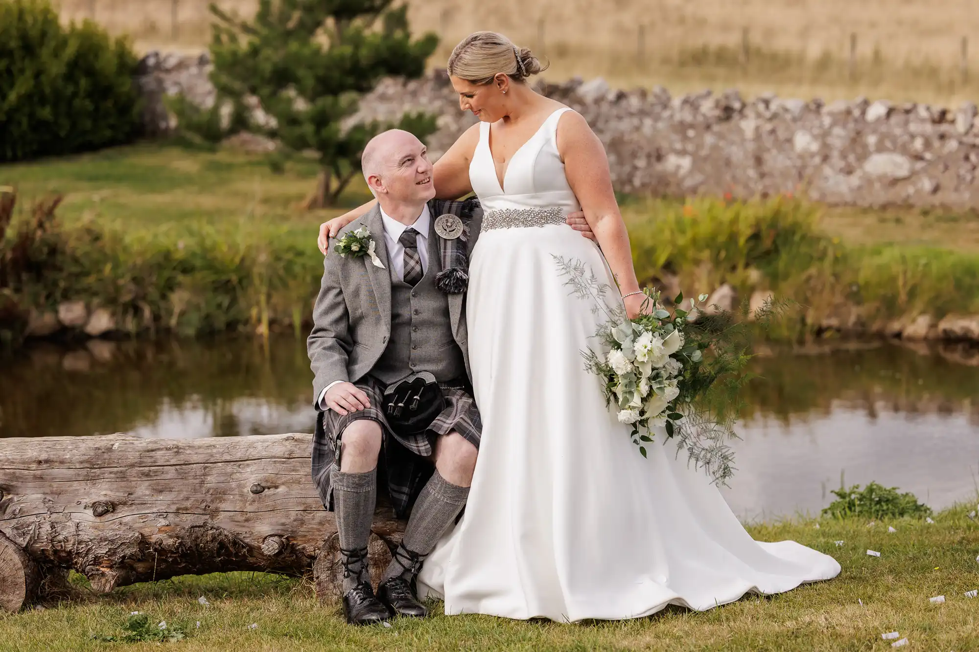 A bride in a white gown holds a bouquet and stands beside a groom in a grey Scottish kilt outfit, seated on a log. They gaze at each other, smiling, with greenery, a pond, and a stone wall in the background.