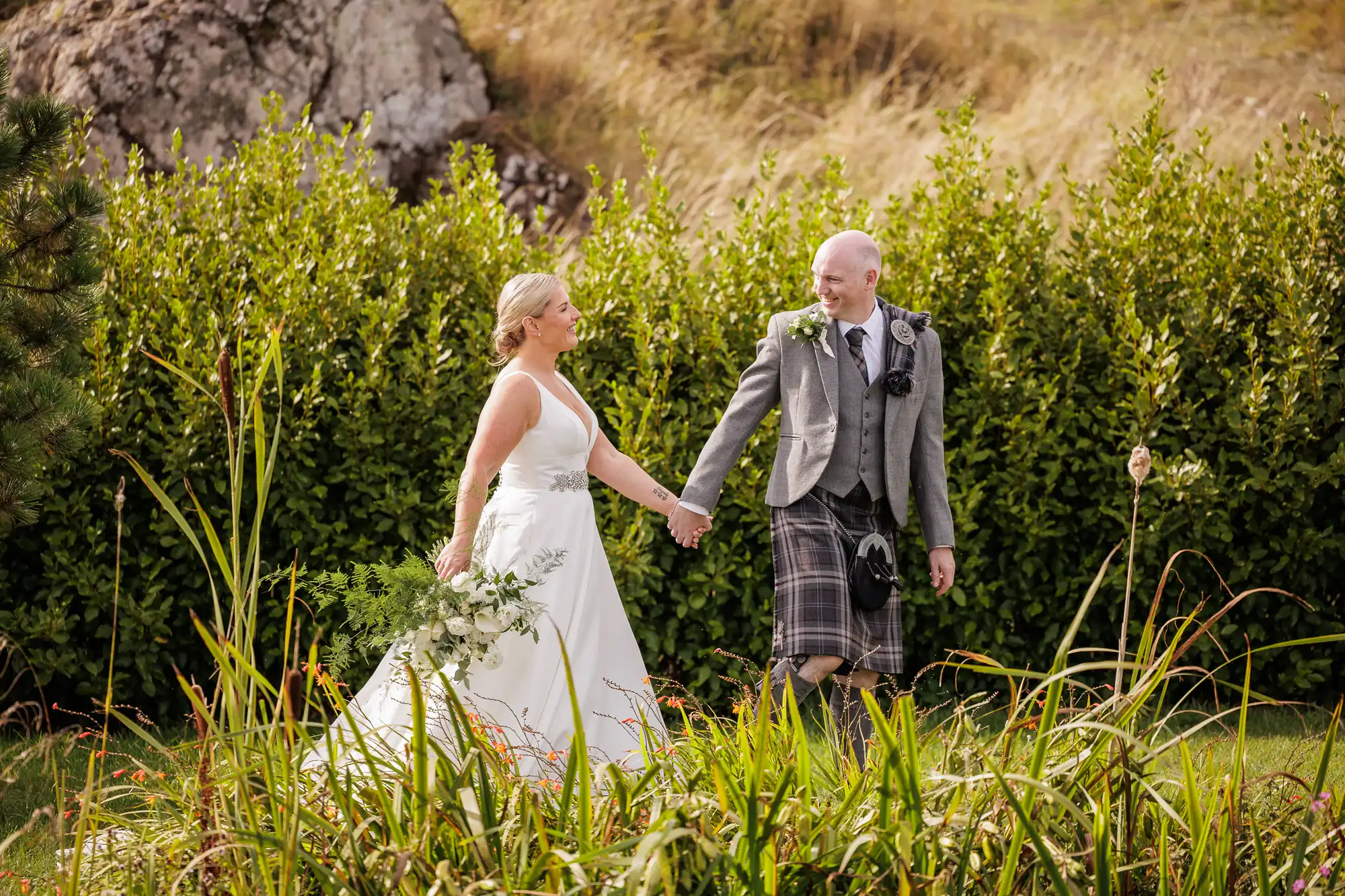 A bride in a white gown and a groom in a grey jacket with a tartan kilt hold hands and smile at each other whilst walking outdoors, surrounded by tall grasses, green bushes, and a rocky hillside in the background.
