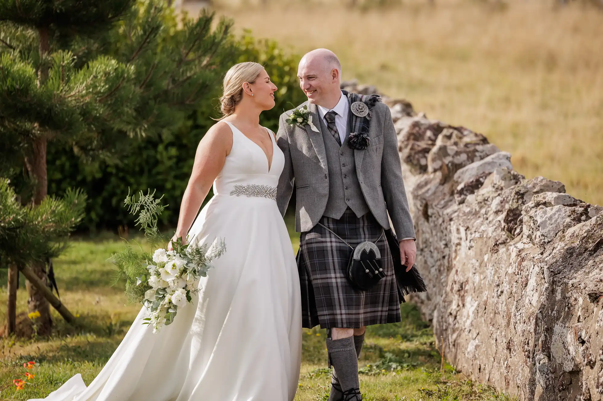 A bride in a white dress holding a bouquet and a groom in a grey jacket and kilt walk outdoors, smiling at each other. They stroll beside a stone wall with grass and greenery in the background, suggesting a countryside wedding setting.
