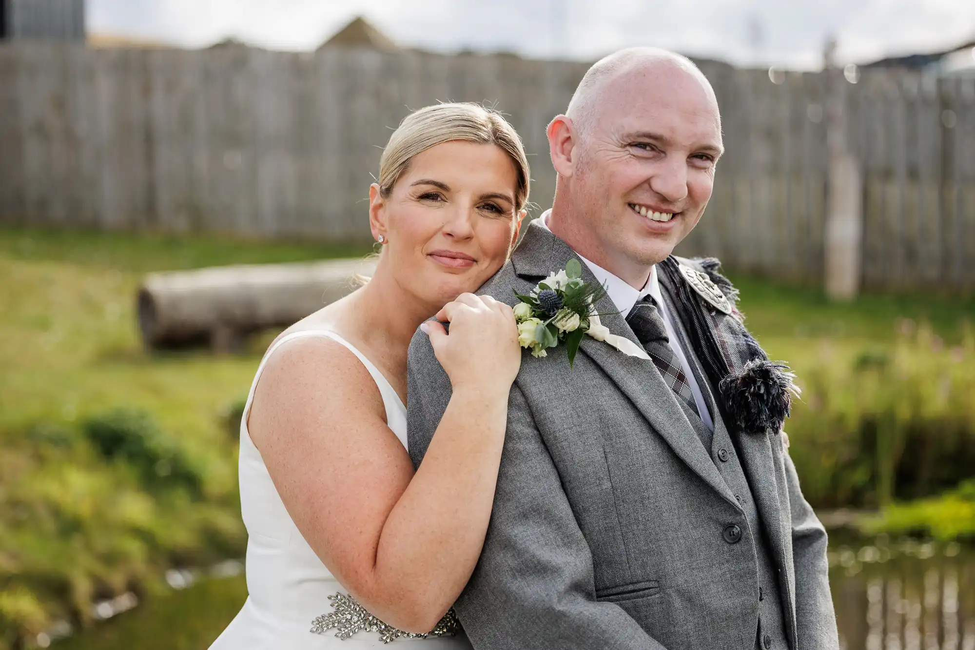 A smiling couple poses outdoors in wedding attire. The woman in a white dress rests her head on the shoulder of the man, who wears a grey suit with a thistle buttonhole. They stand in front of a grassy area with a wooden fence and log in the background.