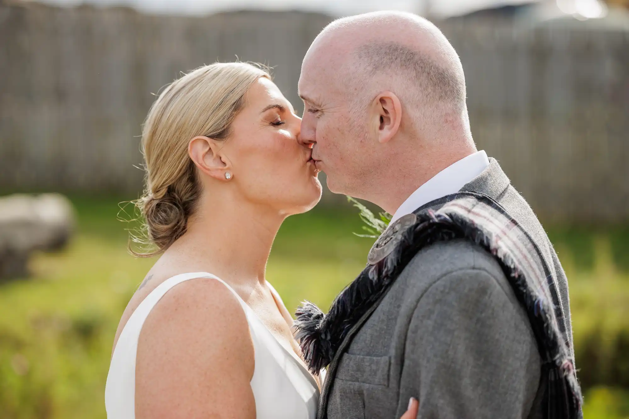 A bride and groom share a kiss outdoors. The bride has blonde hair styled in a low bun and wears a white dress. The groom, bald, wears a grey suit with a patterned sash. A wooden fence and greenery appear blurred in the background.