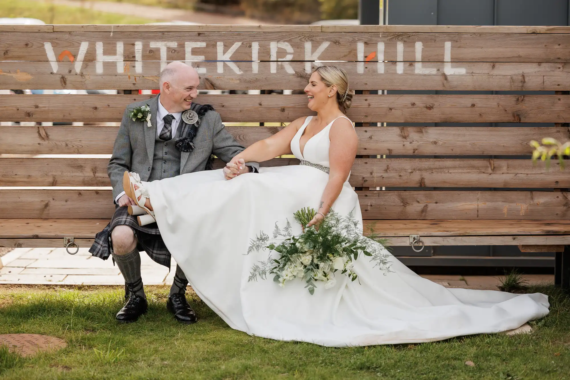 A joyful bride in a white gown and bouquet sits on a wooden bench beside a smiling groom in a grey kilt outfit. They hold hands and laugh beneath a sign reading "WHITEKIRK HILL" outdoors on green grass.