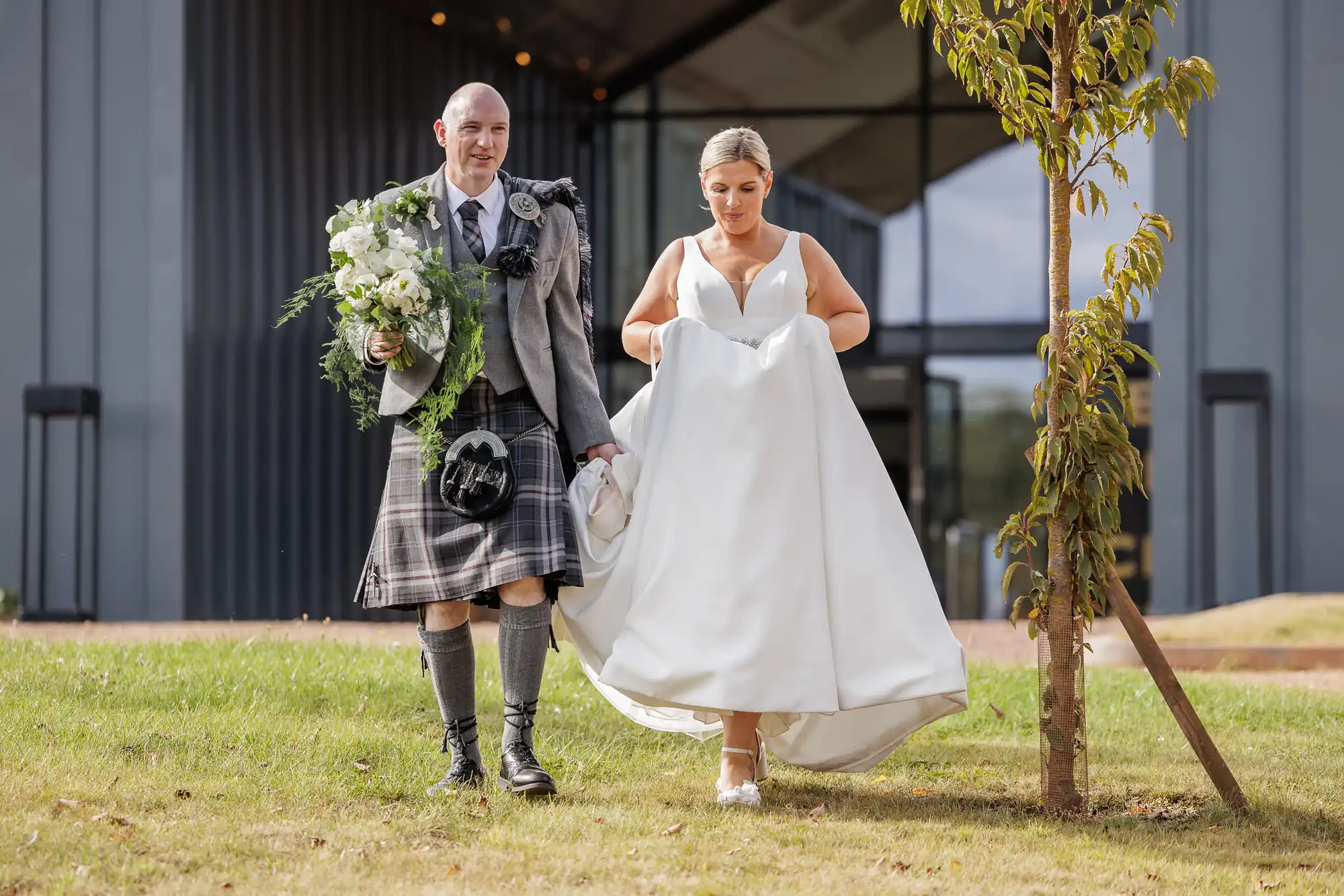 A groom in a grey jacket, kilt, and knee-high socks walks beside a bride in a white sleeveless dress holding her gown. The groom carries a bouquet and both are walking on grass outside a modern building with large glass doors.