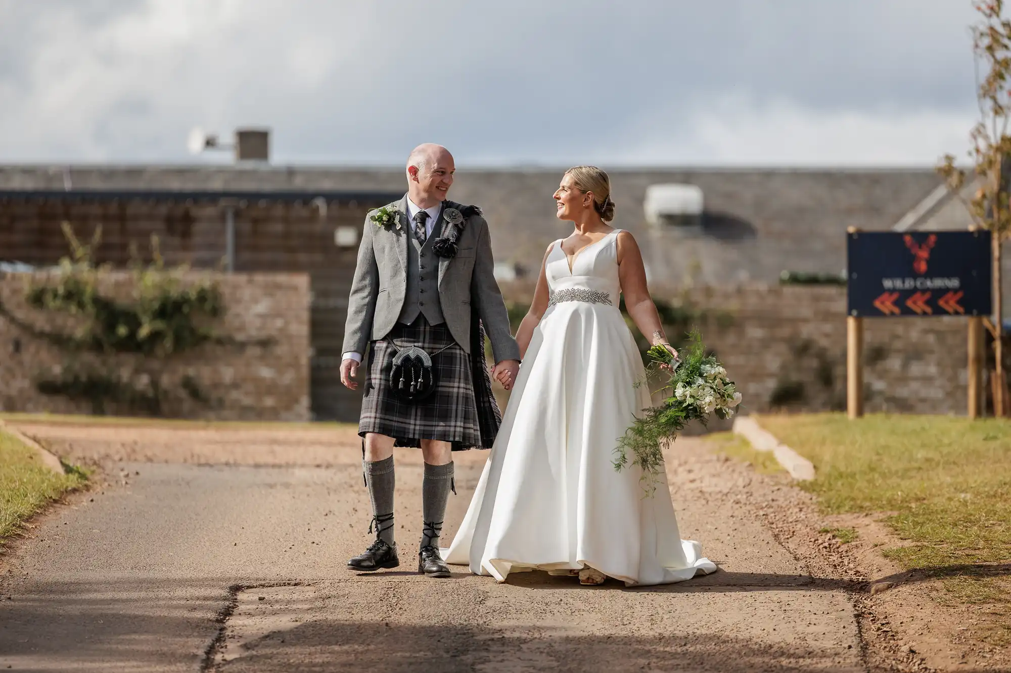 A groom in a kilt and jacket holds hands with a bride in a white dress, both smiling as they walk down an outdoor path. The bride carries a bouquet; they look at each other. In the background is a stone building and a sign reading "WEDDINGS.