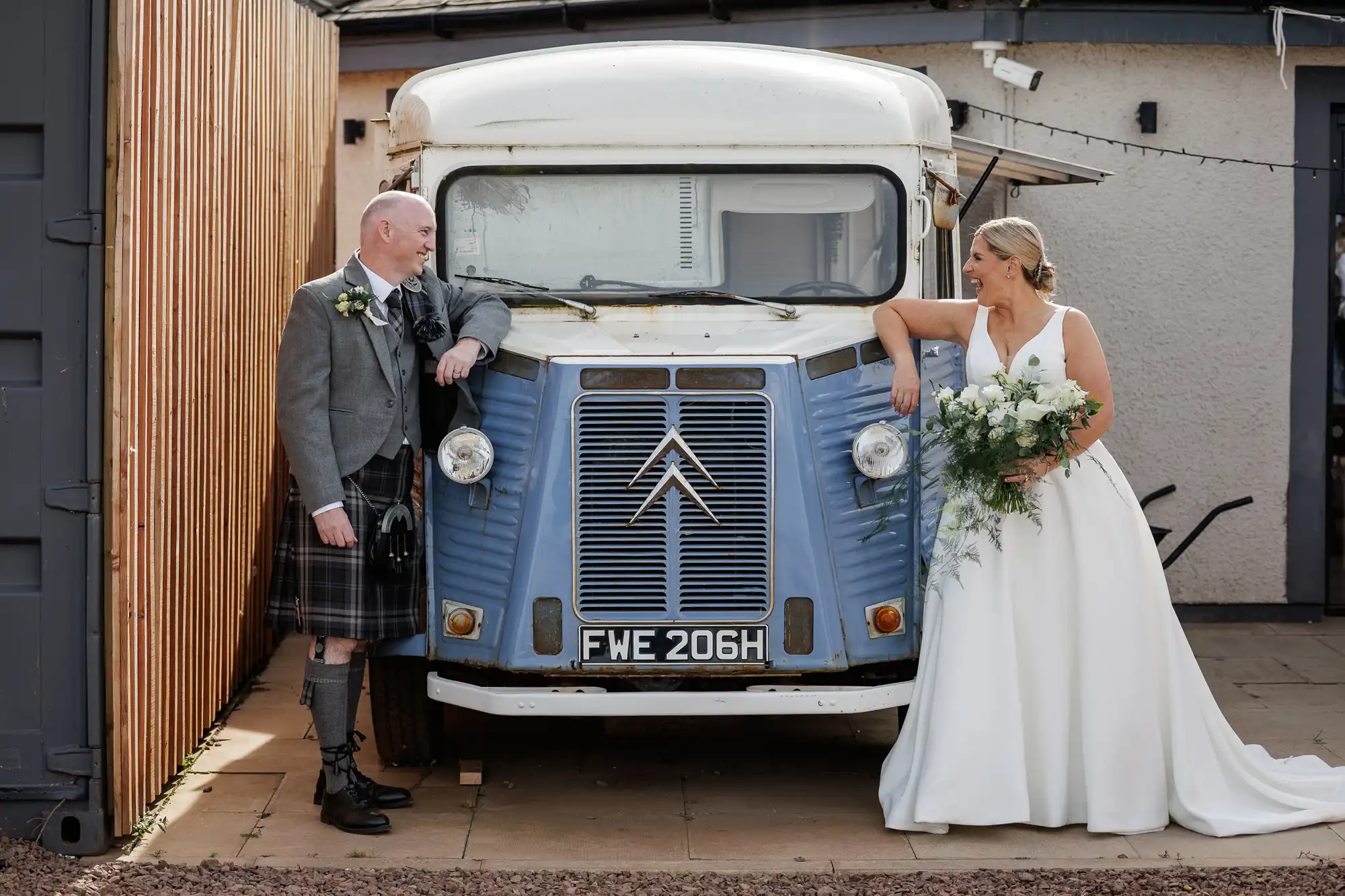 A bride in a white gown holding a bouquet stands beside a groom in a grey jacket and kilt. They smile at each other, leaning on a vintage blue and white Citroën van with the number plate "FWE 206H," parked near a wooden fence and building.