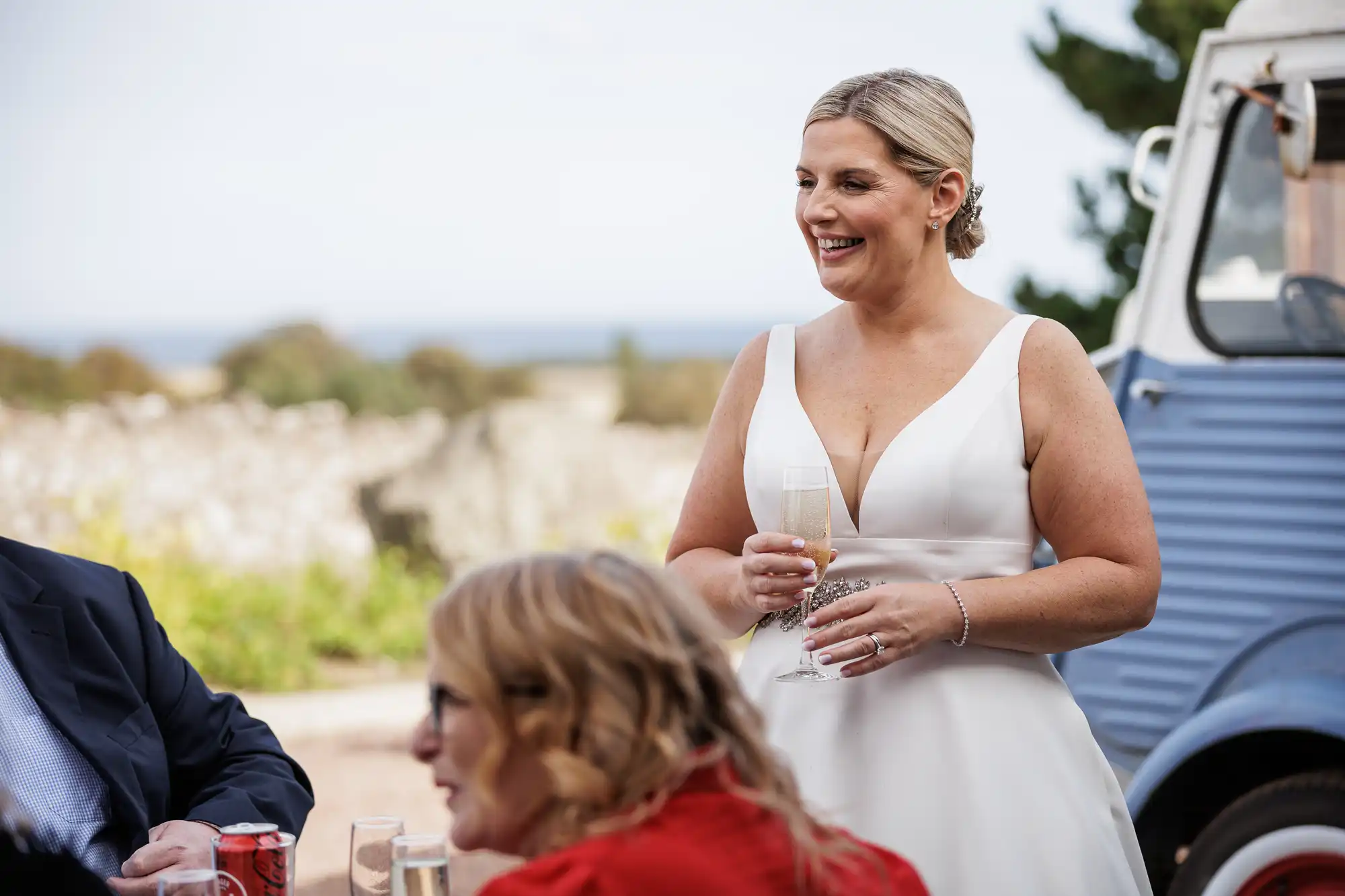 A smiling bride in a white sleeveless dress holds a champagne flute outdoors. She has blonde hair in an updo and wears a bracelet. Guests are seated nearby, and a blue vintage vehicle is in the background with greenery and a stone wall visible.