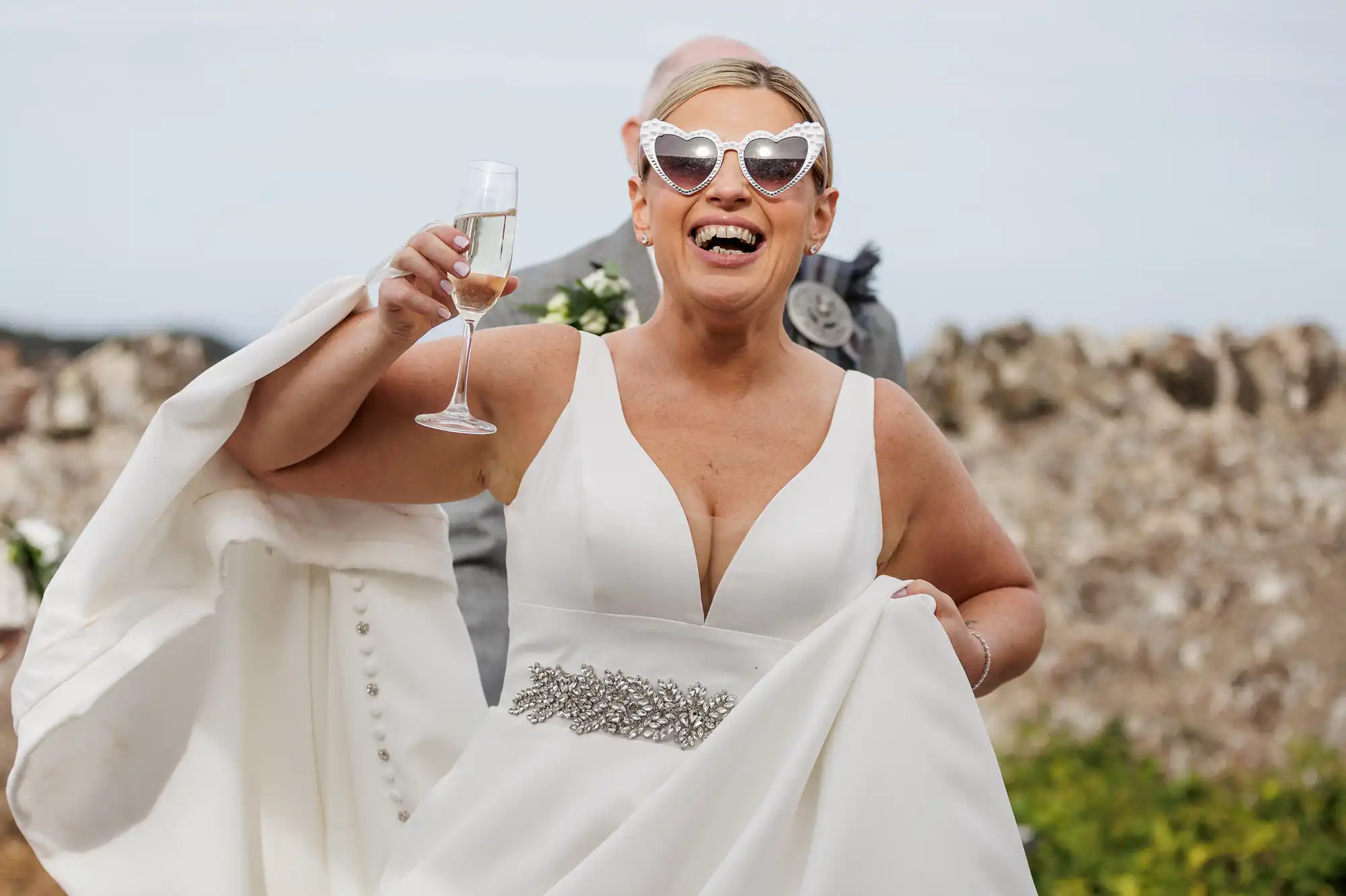 A smiling bride in a white dress and heart-shaped sunglasses holds a glass of champagne and lifts her gown. The background shows an outdoor setting with greenery and a stone wall, and a person partly visible behind her.