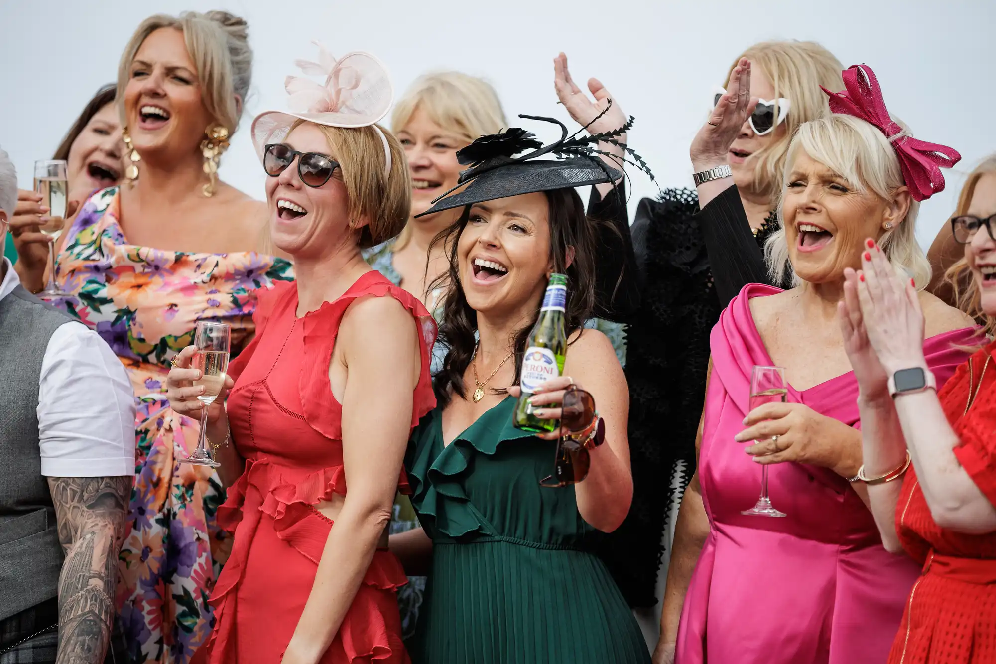 A group of women in colourful dresses and fancy hats smile and cheer together at an outdoor event. Most hold glasses of champagne; one woman in green holds a beer bottle. They appear joyful, celebrating, and dressed for a festive occasion.