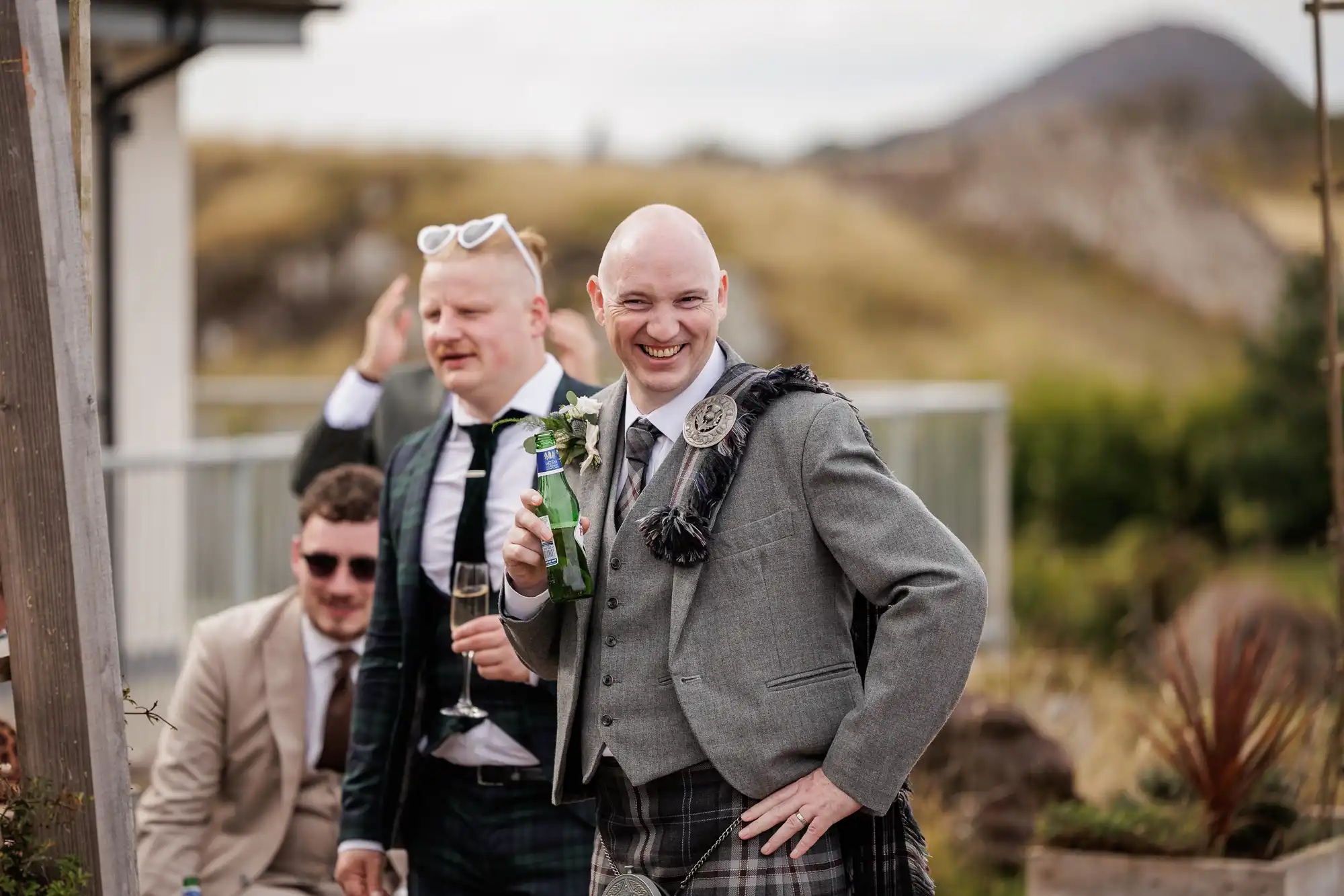 A smiling man in a grey suit and tartan kilt holds a drink and poses outdoors. Two men in suits, one wearing sunglasses on his head, stand behind him. Another man sits. The background shows grassy hills and a mountain under a cloudy sky.