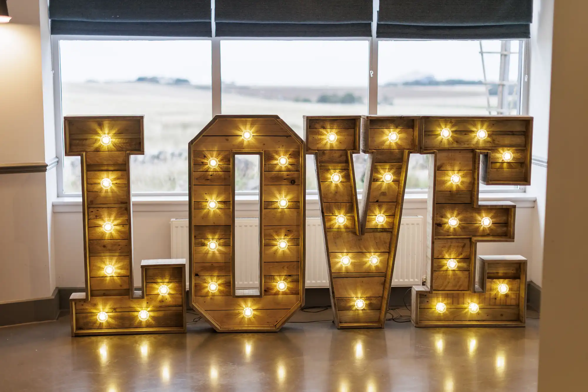 Large wooden letters spelling "LOVE" stand indoors in front of a window. Each letter is lit up with round, exposed bulbs, creating a warm, glowing effect. The floor is shiny, and there is natural light coming from the window behind.