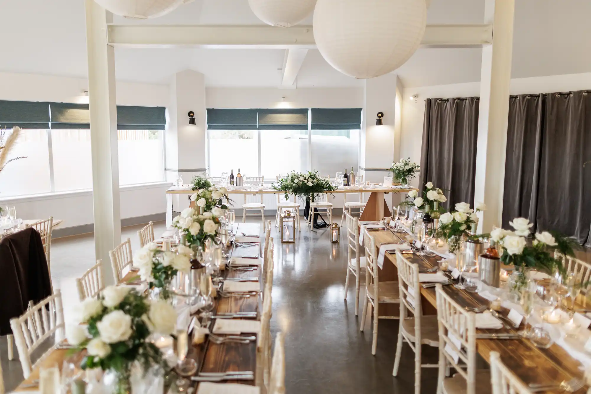 A bright, elegant dining room set for an event, with wooden tables, white chairs, and white floral centrepieces. White serviettes and glassware are neatly arranged, and paper lanterns hang from the ceiling. Large windows let in natural light.