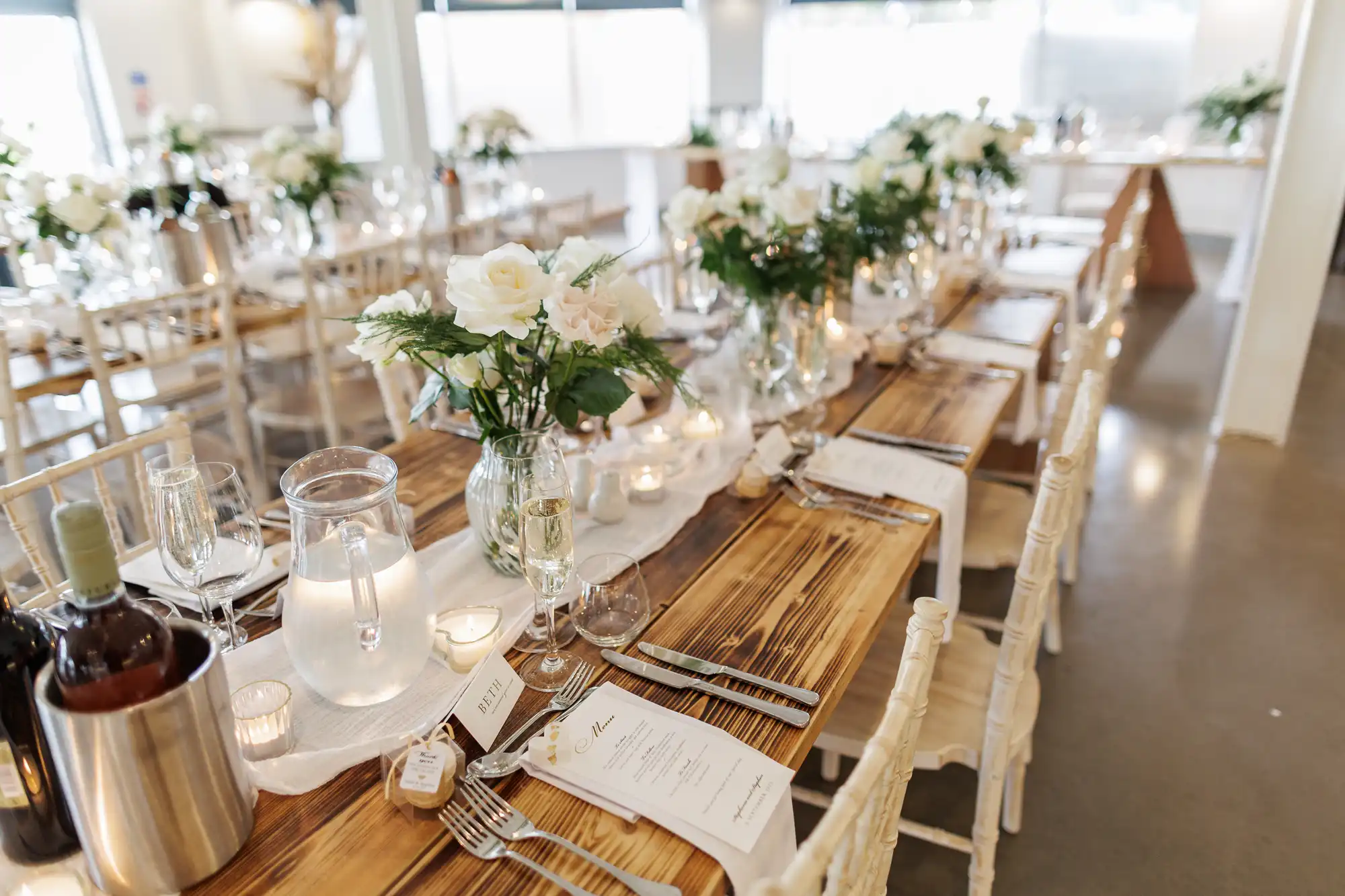 A wooden table set for a formal event with white floral centrepieces, wine, water jugs, glassware, menus, and candles. Ivory chairs line both sides of the table in a bright, elegant room with large windows in the background.