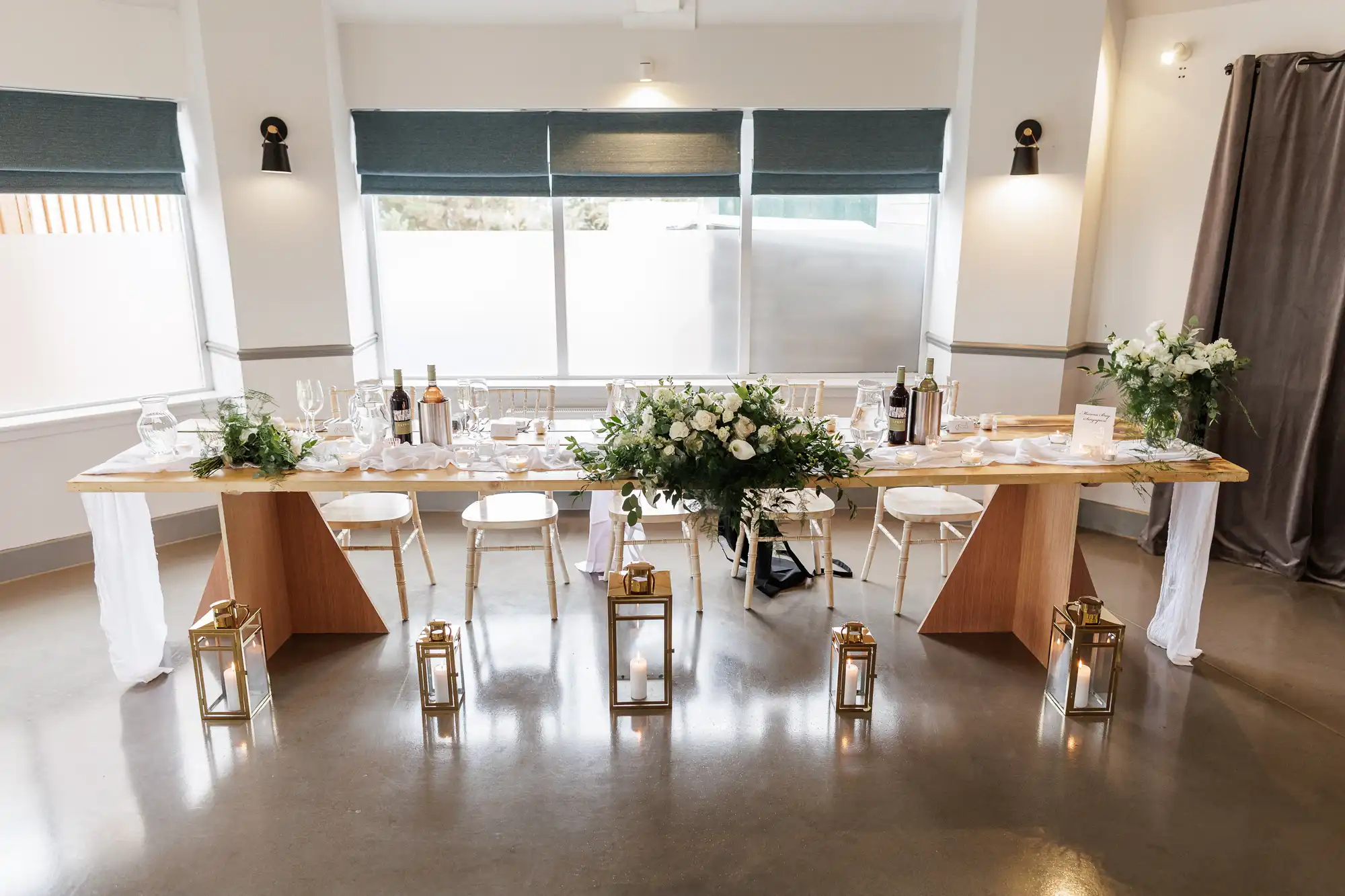 A long wooden table set for a formal event is adorned with white plates, wine glasses, candles, bottles, and a centrepiece of white flowers and greenery. Gold lanterns with candles are arranged on the floor in front of the table. White chairs line both sides.