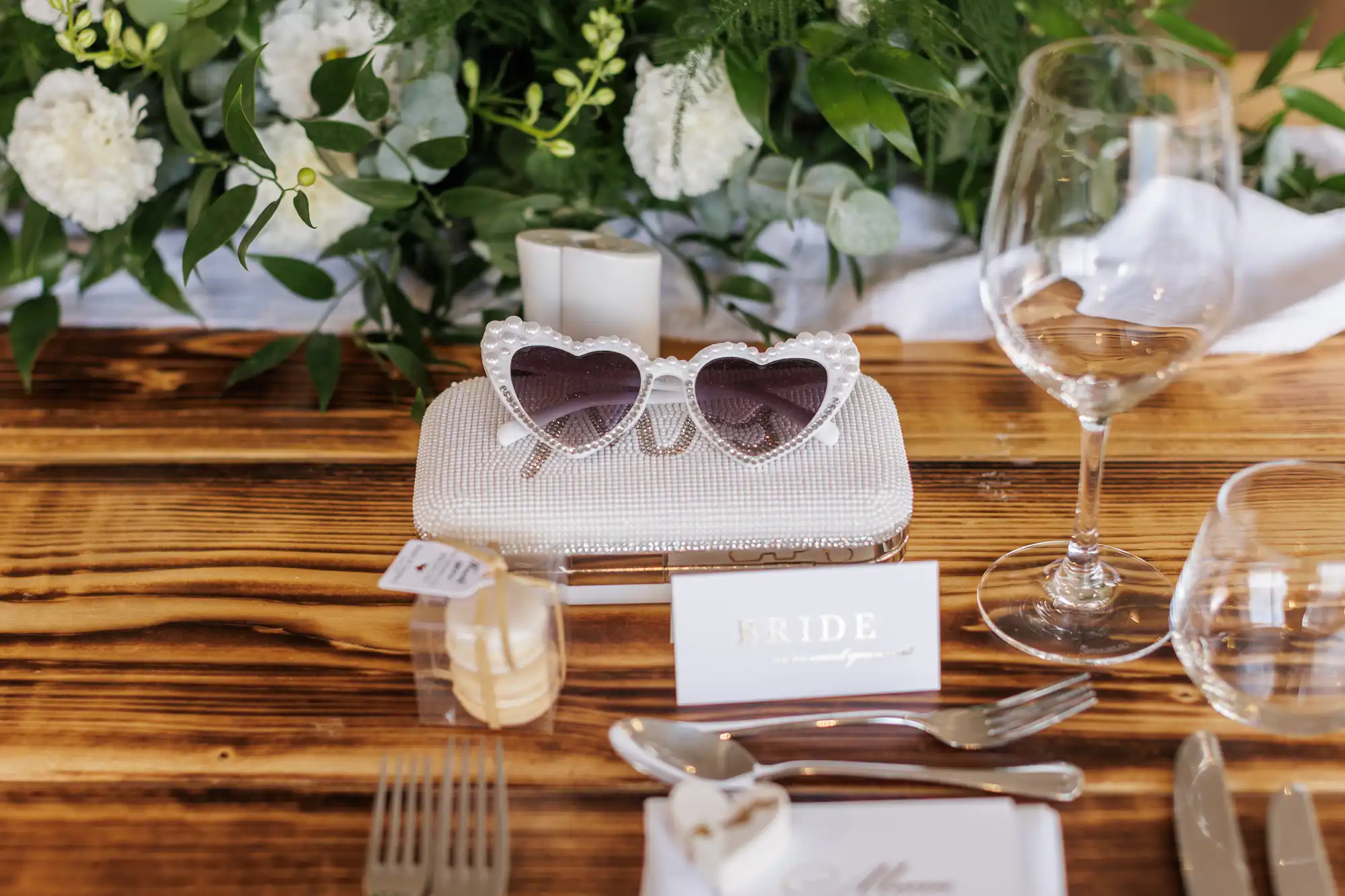 A wooden table set for a wedding, featuring a beaded clutch, heart-shaped sunglasses, a "BRIDE" name card, wine glasses, cutlery, a candle, and lush green and white floral centrepiece in the background.