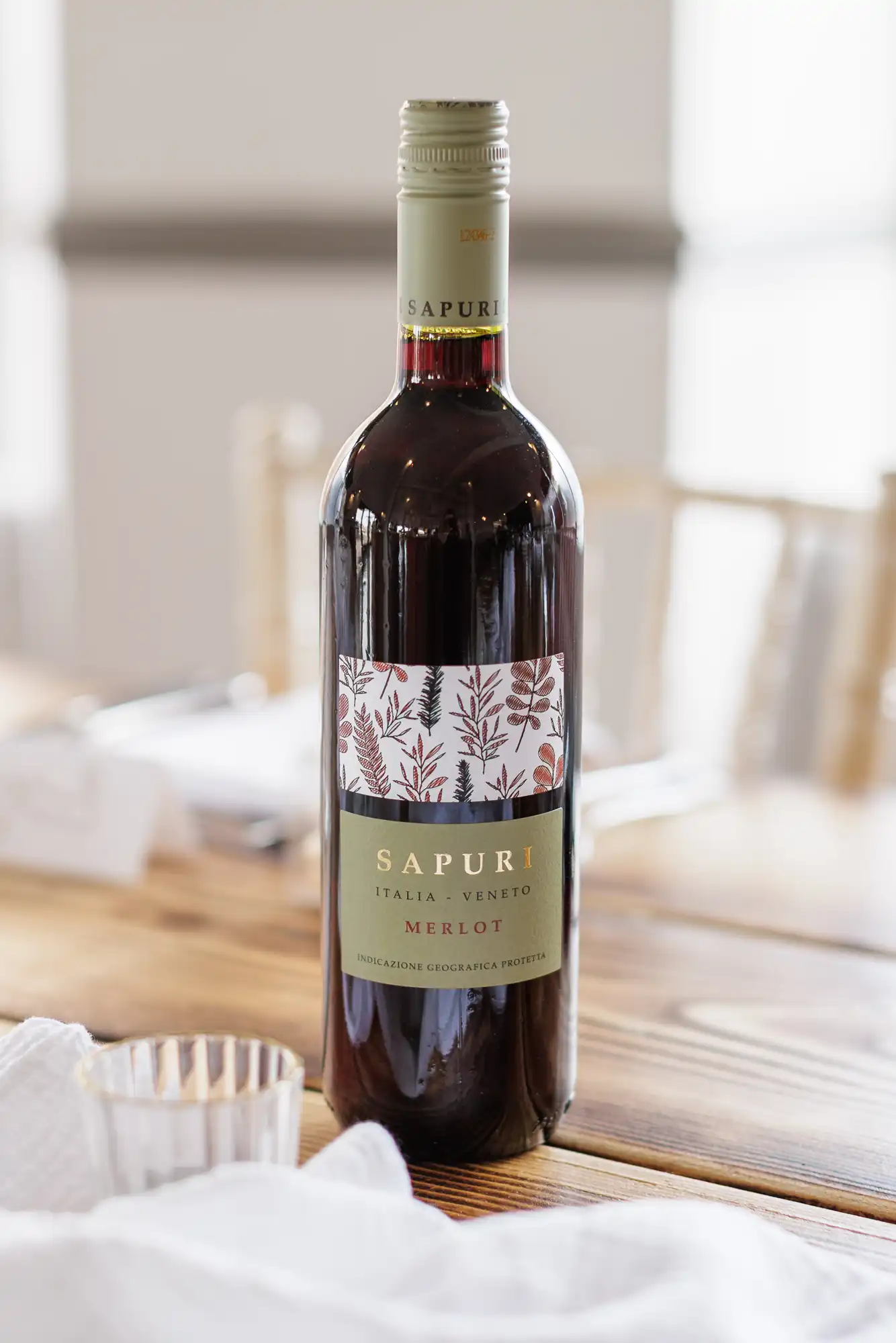 A bottle of Sapuri Merlot wine sits on a wooden table, with a decorative label featuring leaves and twigs. The wine is deep red. A clear, empty glass and a white serviette are nearby, with blurred chairs and crockery in the background.