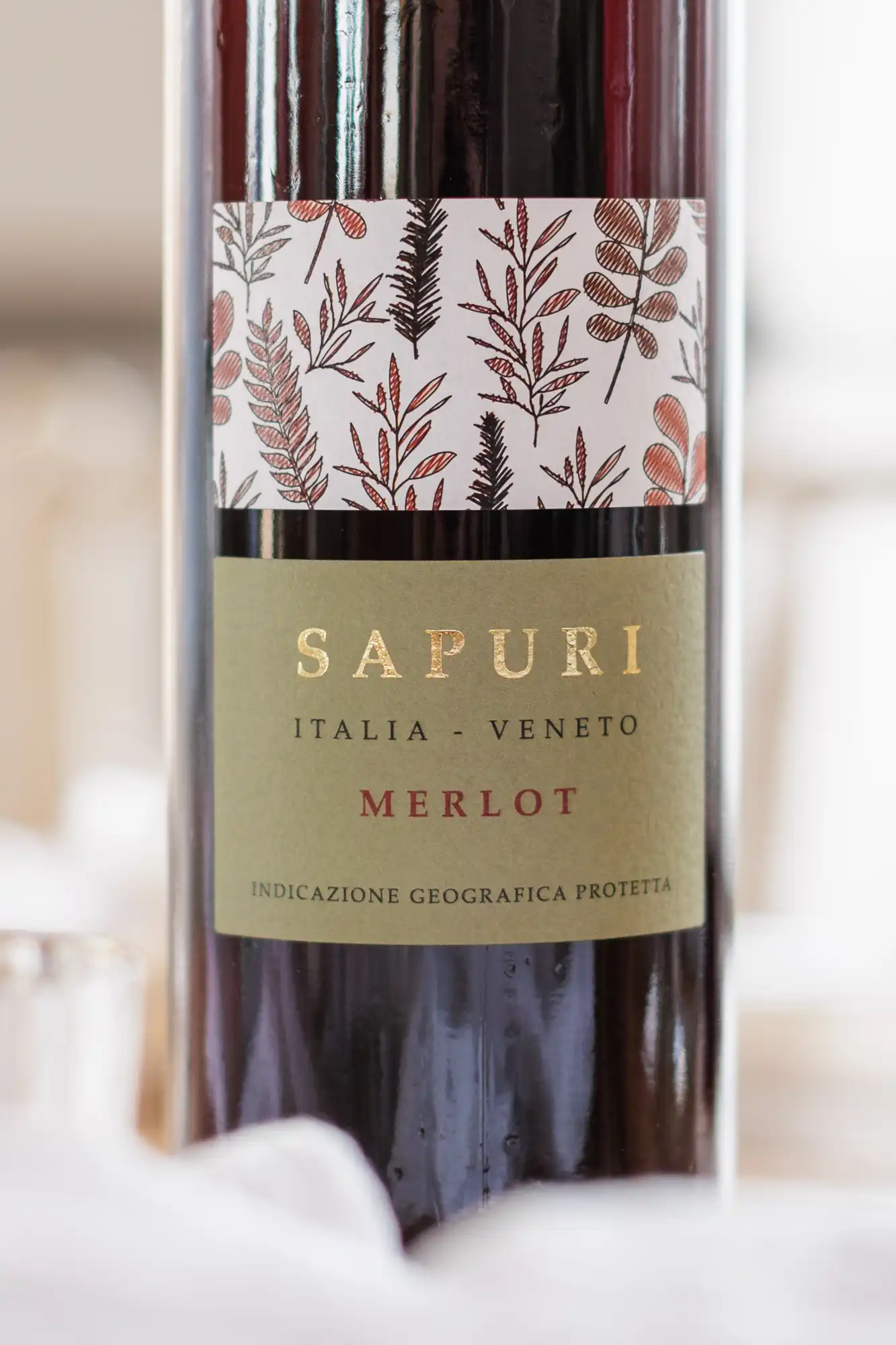 A close-up of a wine bottle labelled "SAPURI" with "ITALIA - VENETO" and "MERLOT" below. The label features a gold background with elegant serif text and a decorative band of illustrated brown and red leaves above the main label.