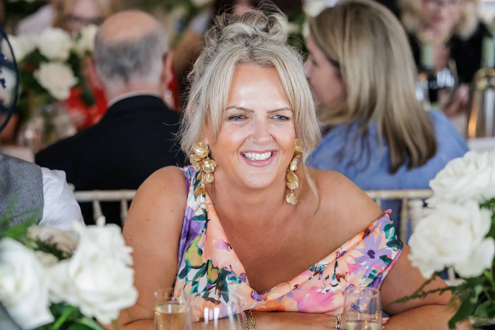 A smiling woman with blonde hair in an updo wears a colourful floral dress and gold earrings, seated at a table decorated with white roses and surrounded by other people in a festive indoor setting.