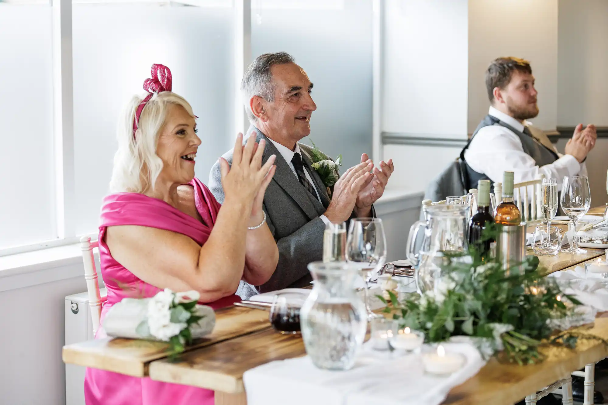 An older woman in a bright pink dress and fascinator and an older man in a grey suit sit at a decorated wooden table, clapping and smiling. A younger man in a waistcoat also applauds. The table has glasses, bottles, greenery, and candles.