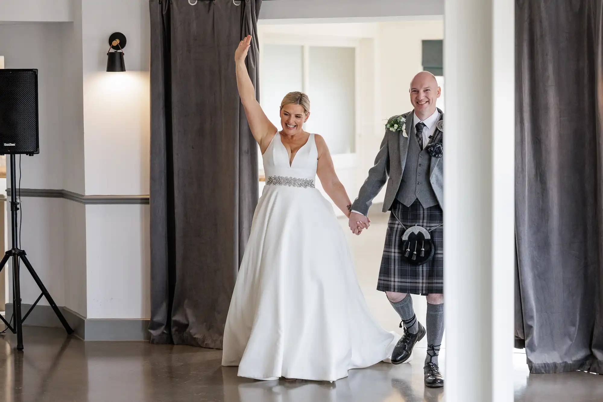 A smiling bride in a white dress raises her arm joyfully while holding hands with a groom in a grey jacket and checked kilt. They are entering a bright room with grey curtains, suggesting a wedding celebration or reception.
