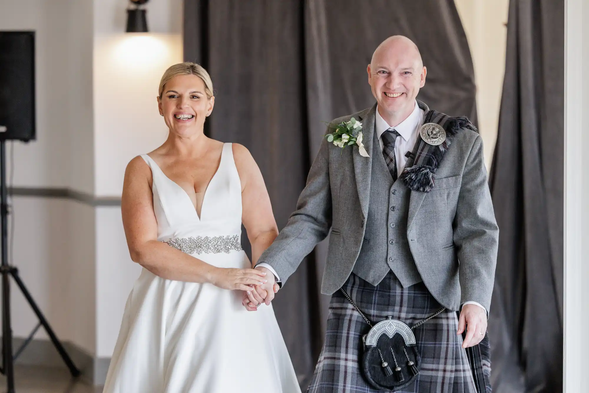 A smiling bride in a white sleeveless gown holds hands with a groom in a grey jacket, tartan kilt, and sporran. Both stand indoors, the groom wearing a buttonhole, with neutral walls and dark curtains in the background.