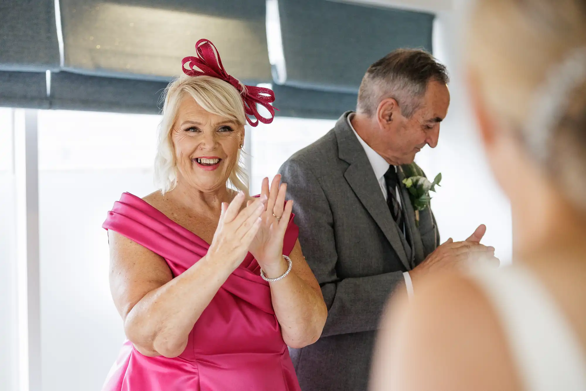 A woman in a bright pink dress and matching fascinator smiles and claps, looking at a person in white (foreground, blurred). Beside her stands a man in a grey suit with a buttonhole, also clapping. Both appear joyful in a well-lit room.