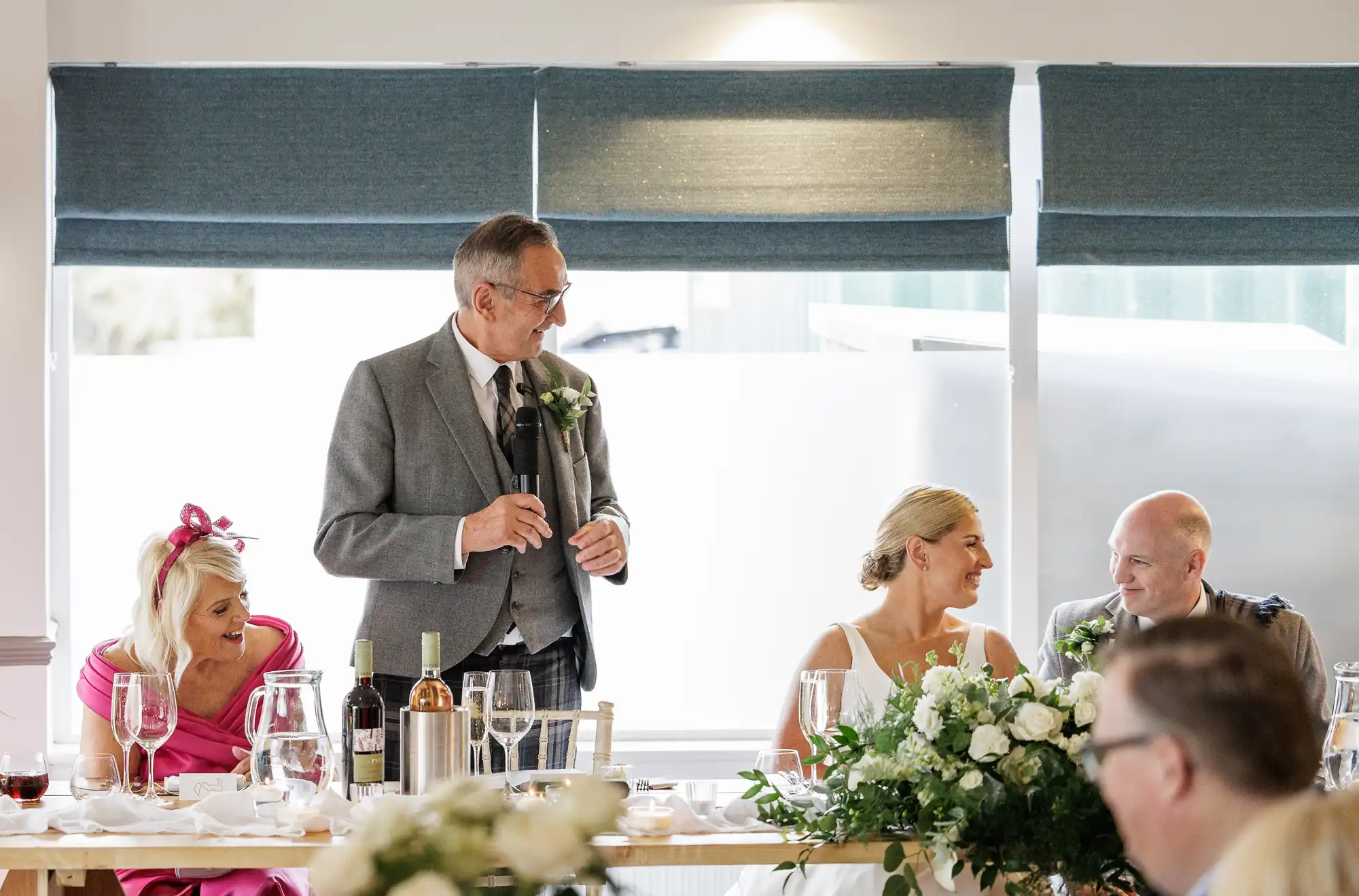 A man in a grey suit stands giving a speech at a wedding reception table, holding a microphone. A bride and groom sit smiling at each other beside him. Guests dressed formally sit on either side. The table is decorated with white flowers and bottles of wine.