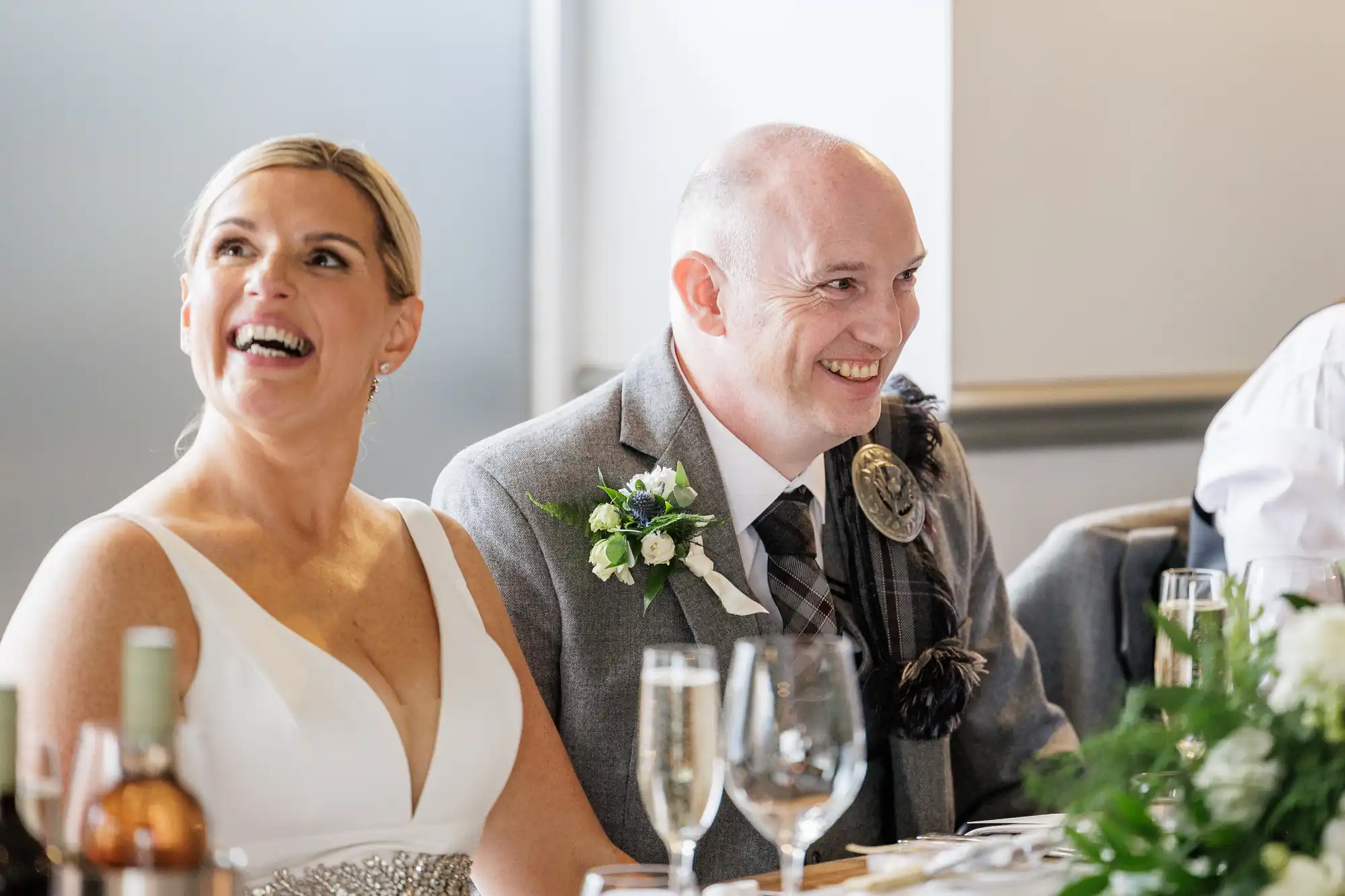 A smiling bride in a white dress and a groom in a grey suit with a buttonhole sit together at a table, enjoying their wedding reception. Glasses, a wine bottle, and green floral arrangements are visible in the foreground.