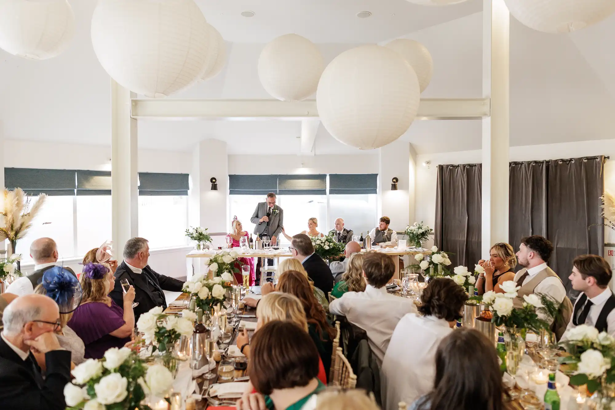 A wedding reception scene with guests seated at decorated tables, listening to a person giving a speech at the top table. White flowers and candles decorate the tables; large white paper lanterns hang from the ceiling. The atmosphere appears joyful and festive.