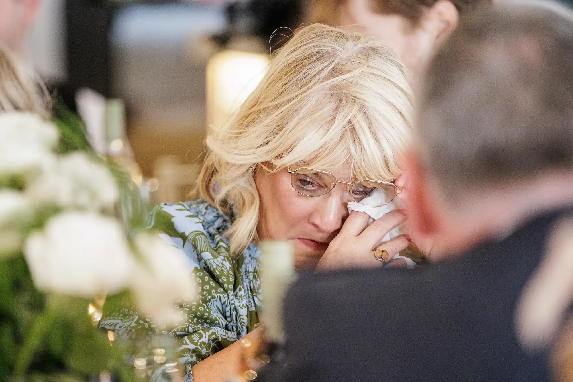A woman with blonde hair and glasses wipes a tear from her eye with a tissue at a table, surrounded by blurred people and white flowers, suggesting an emotional moment at a social gathering or event.