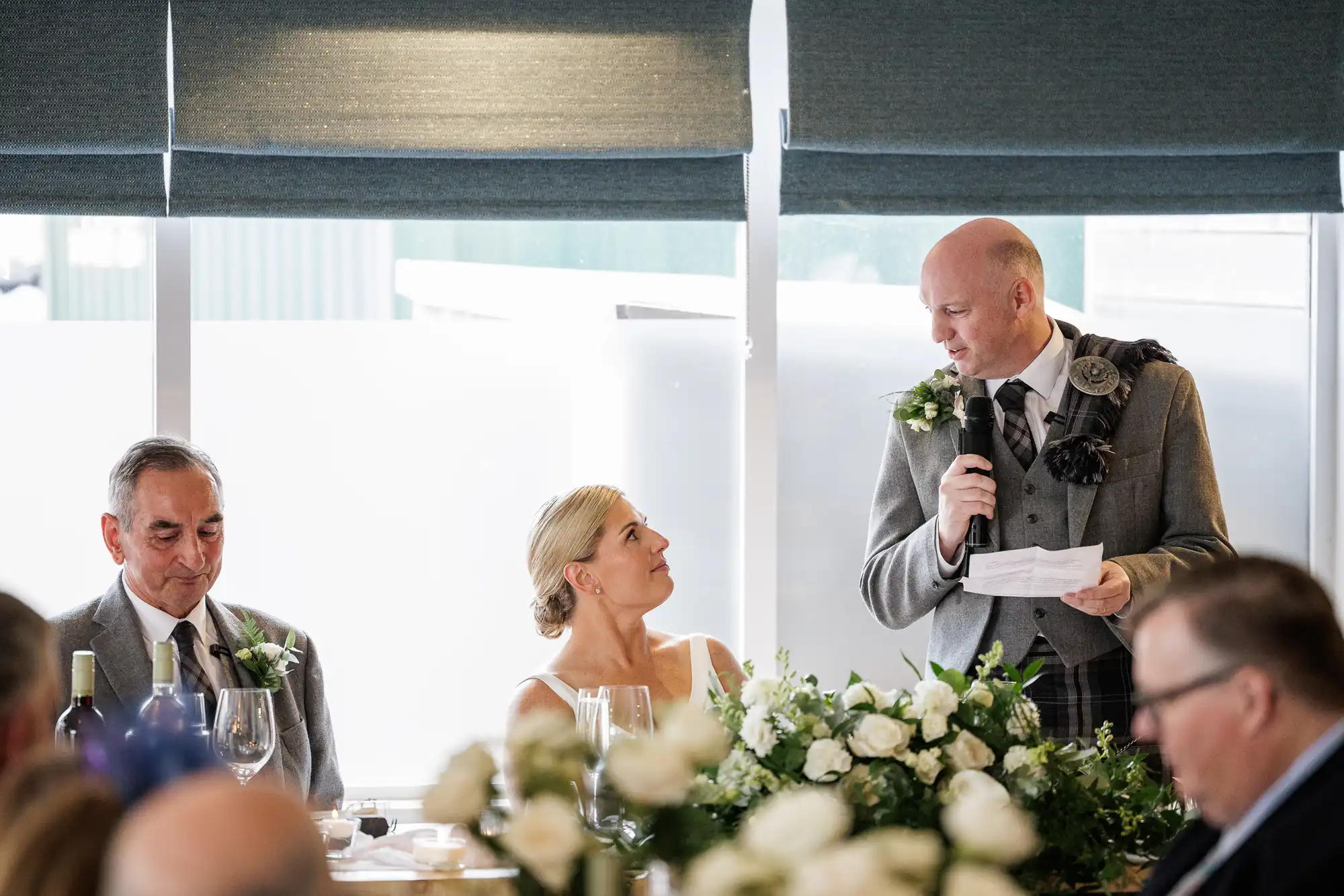 A bride in a white dress sits at a table with white flowers, looking up at a man in a grey suit giving a speech with a microphone and paper. Another man, also in grey, sits on her other side. Guests listen in the foreground. Large windows are behind them.