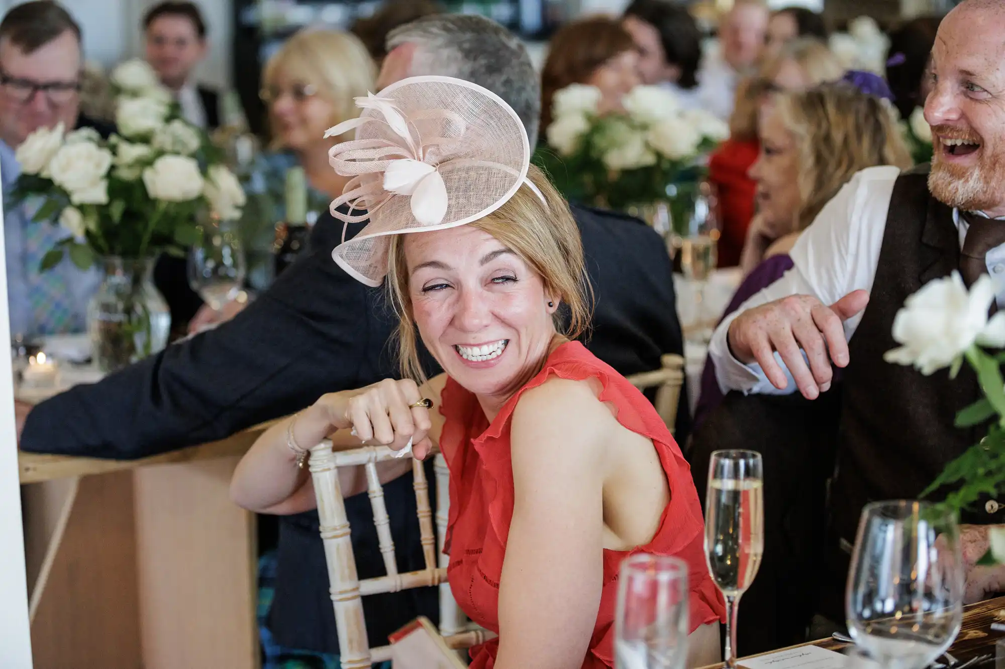 A woman in a red dress and pink fascinator laughs heartily at a crowded event. She sits at a table with white roses and glasses of champagne, surrounded by smiling, well-dressed guests, suggesting a lively, celebratory atmosphere, likely a wedding or party.