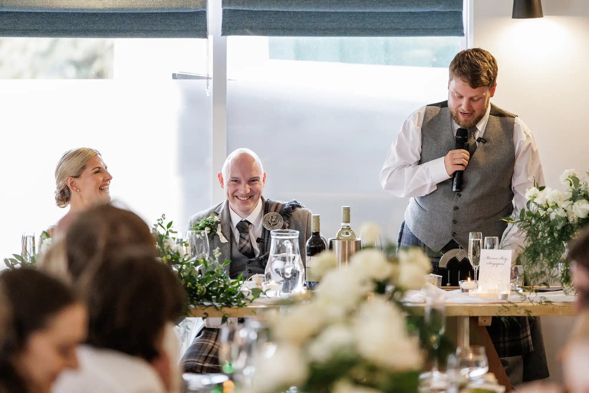 A man in a grey waistcoat speaks into a microphone at a decorated table, smiling guests beside him. The table is adorned with greenery, bottles, and glasses. Natural light filters through windows behind, and blurred floral arrangements are in the foreground.