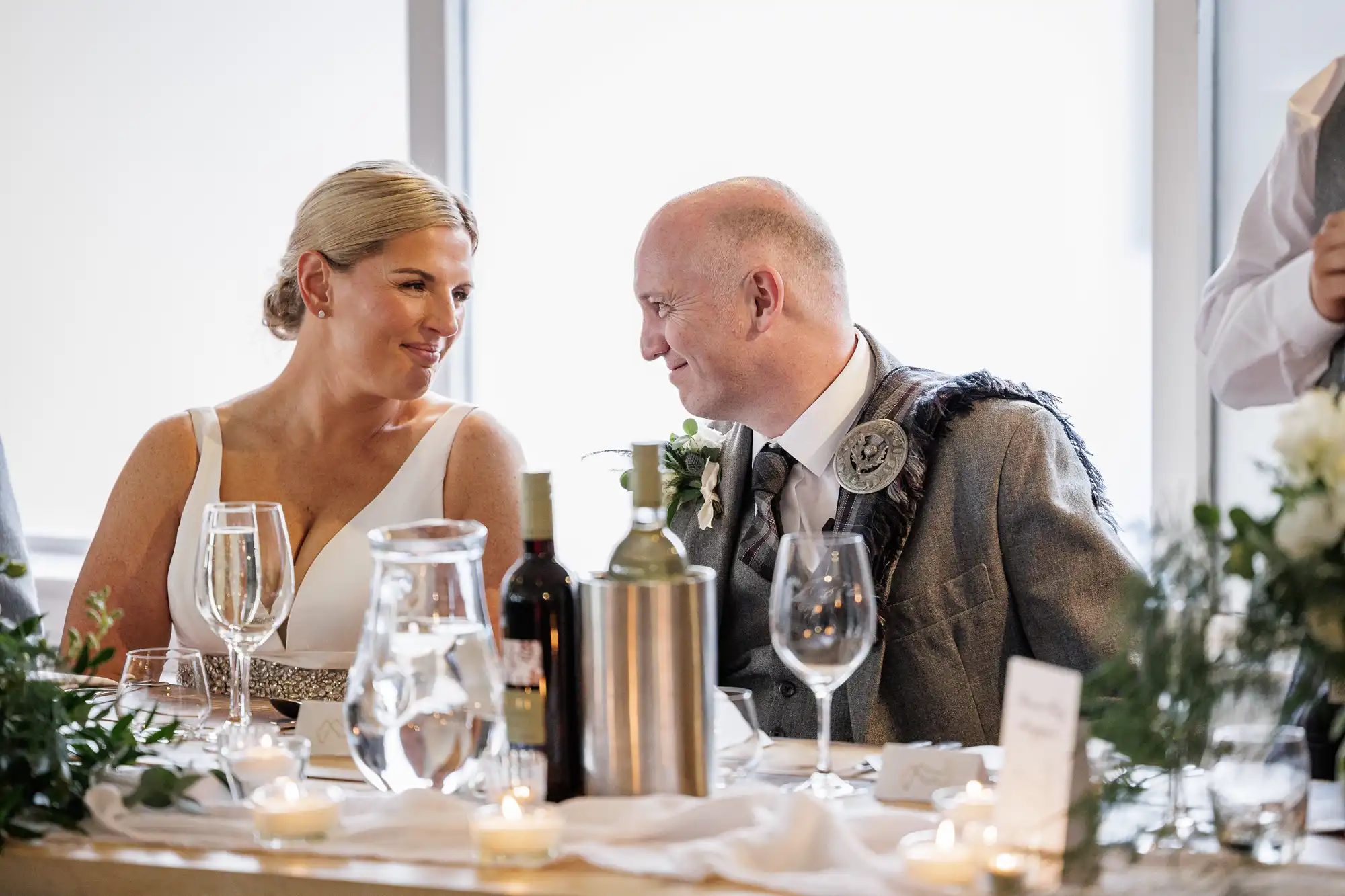 A bride in a white dress and a man in a grey kilt jacket sit at a decorated table, smiling warmly at each other. The table has wine bottles, glasses, candles, greenery, and a white tablecloth. Bright natural light fills the background.