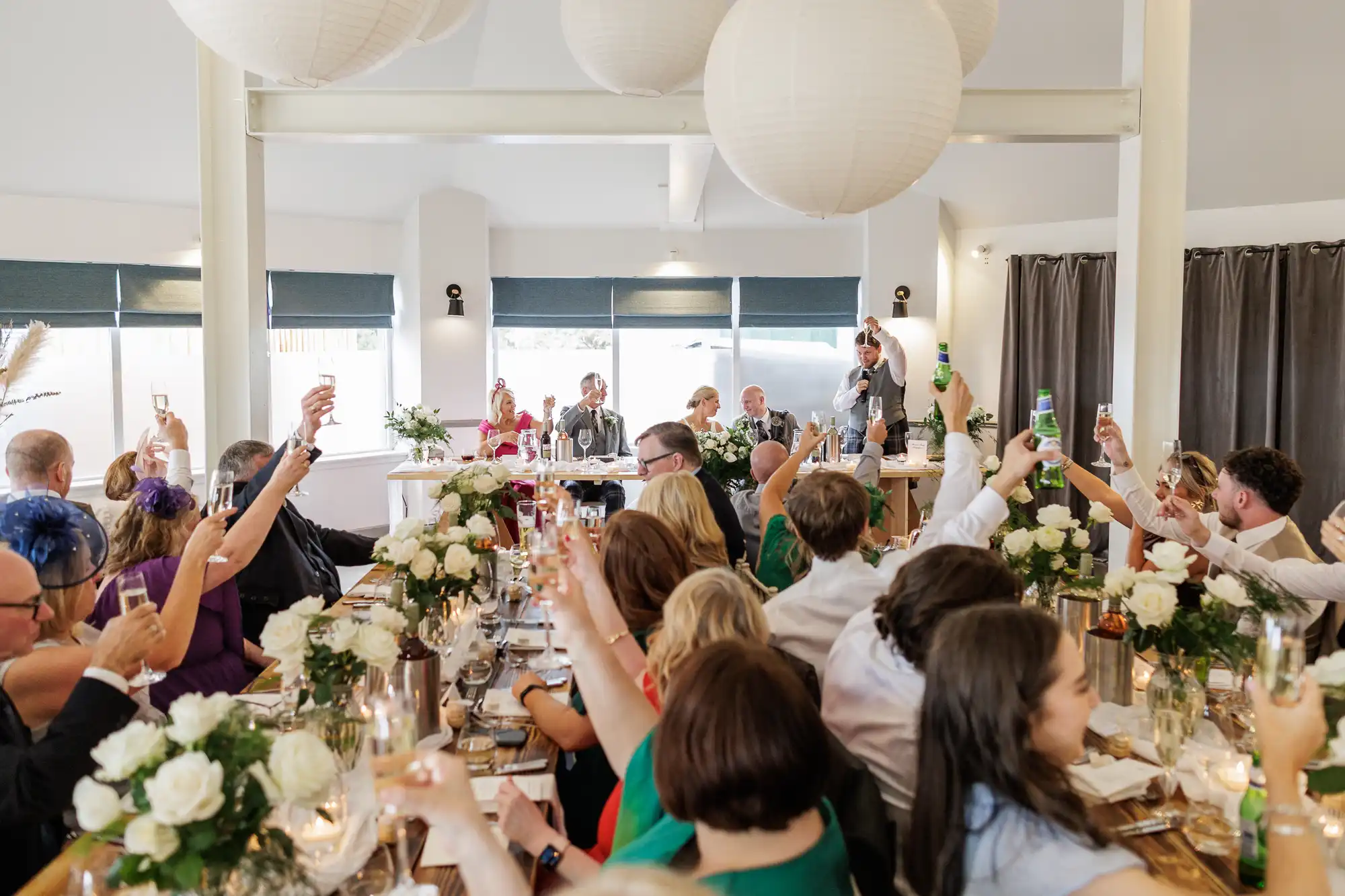 Wedding guests seated at decorated tables raise glasses in a toast towards a speaker at the head of the room. The setting features white flowers, candles, and large hanging paper lanterns, creating a festive and elegant atmosphere.
