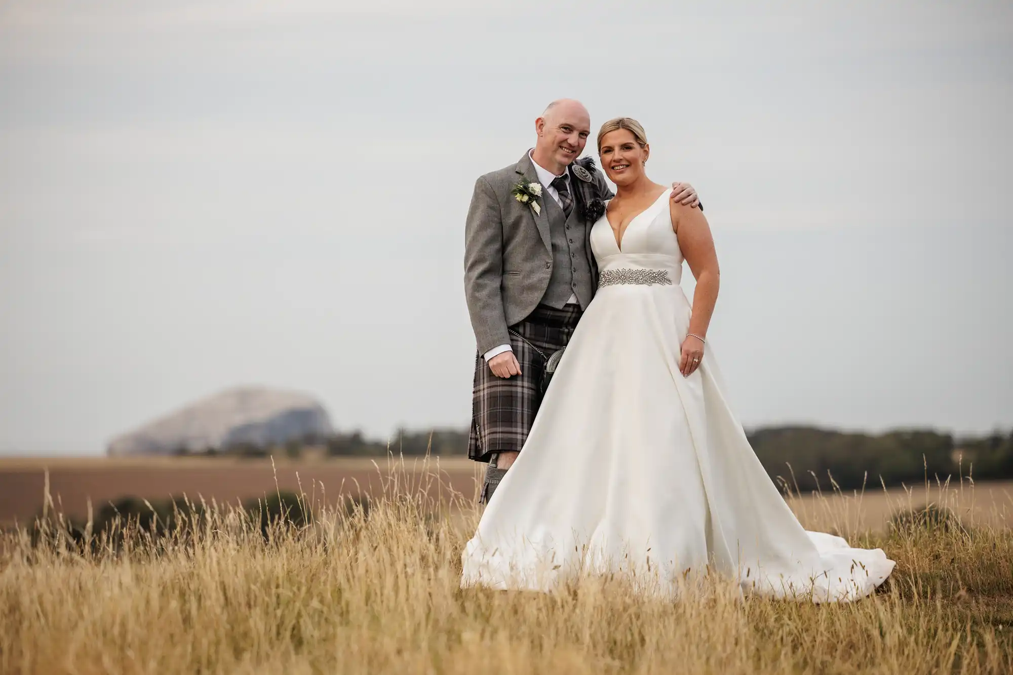 A smiling bride in a white gown stands beside a groom in a grey jacket and kilt. They pose together in a grassy field, with the groom's arm round the bride. A distant rocky hill and cloudy sky can be seen in the background.