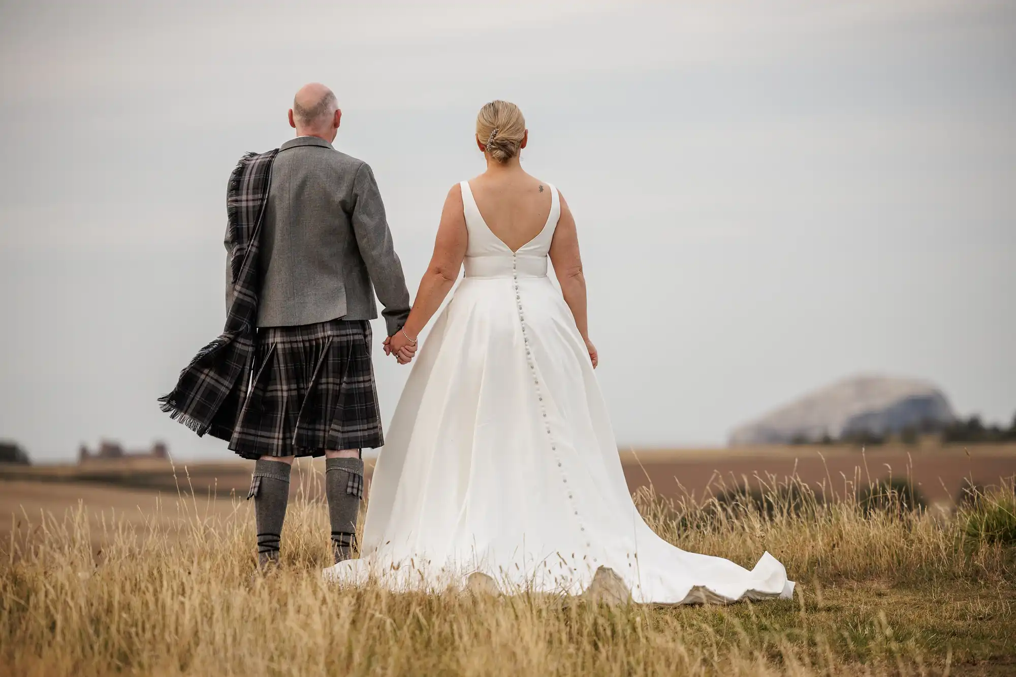 A bride in a white dress and a groom in a kilt stand hand in hand, backs to the camera, in a grassy field. They look towards a distant rock formation under a cloudy sky, evoking a peaceful and romantic mood.