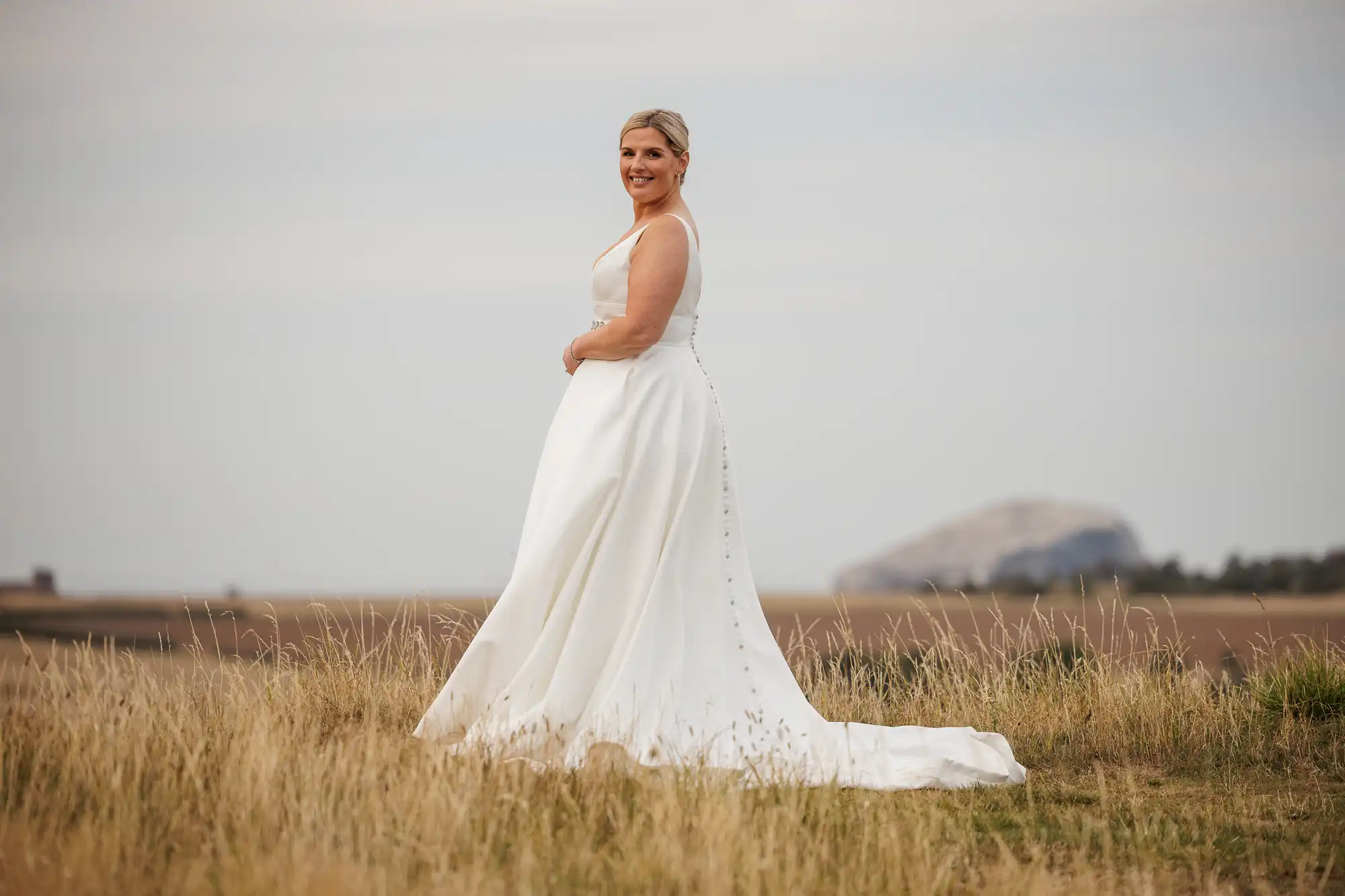 A woman in a white sleeveless wedding dress stands in a grassy field, smiling and looking at the camera. The dress has a long train and button details down the back. The background features open fields and a distant rocky hill under a cloudy sky.