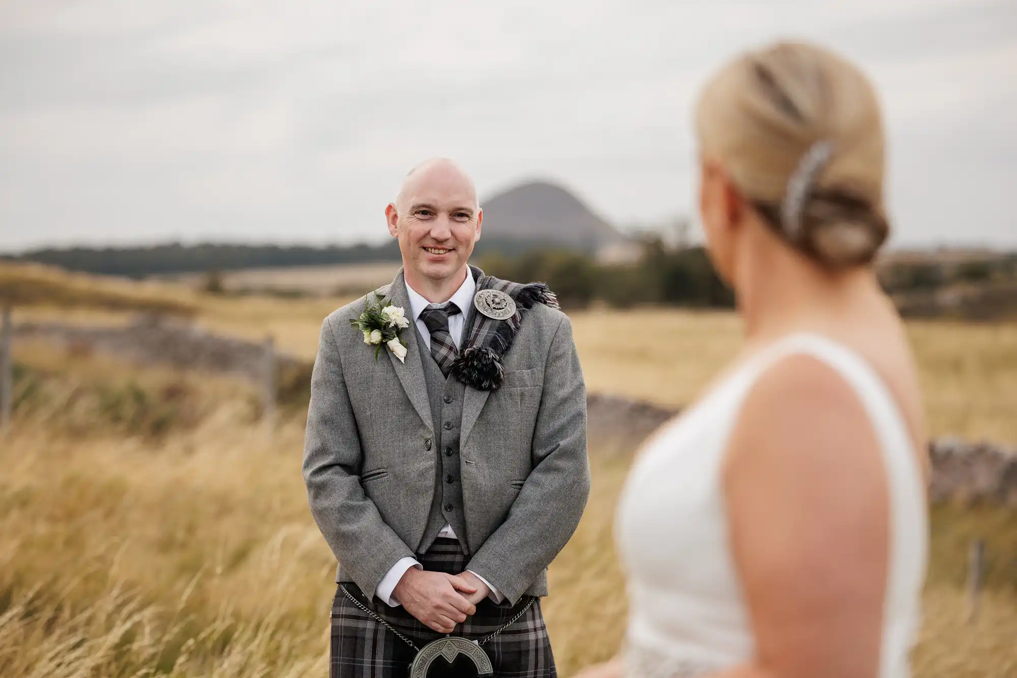 A smiling groom in a grey jacket, flower buttonhole, and tartan kilt stands in a grassy field facing a bride in a white dress; her back is to the camera. A distant hill and stone wall are visible under a cloudy sky.