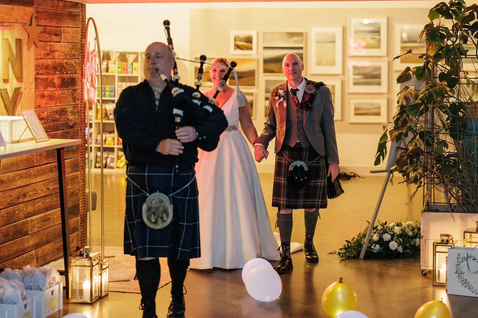 A bagpiper in traditional Scottish attire leads a bride in a white dress and a groom in a kilt through a warmly lit, modern venue decorated with balloons, candles, plants, and framed photos on the walls. The couple hold hands and smile.