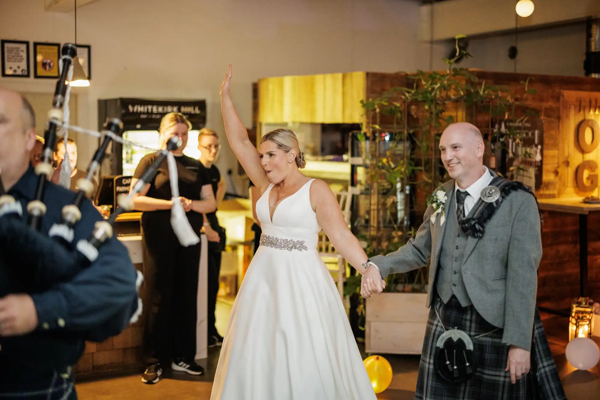 A bride in a white dress with one arm raised and a groom in a kilt hold hands, entering a lively indoor venue. People in the background watch, some in black attire. A bagpiper plays, and the space is decorated with balloons and warm lighting.