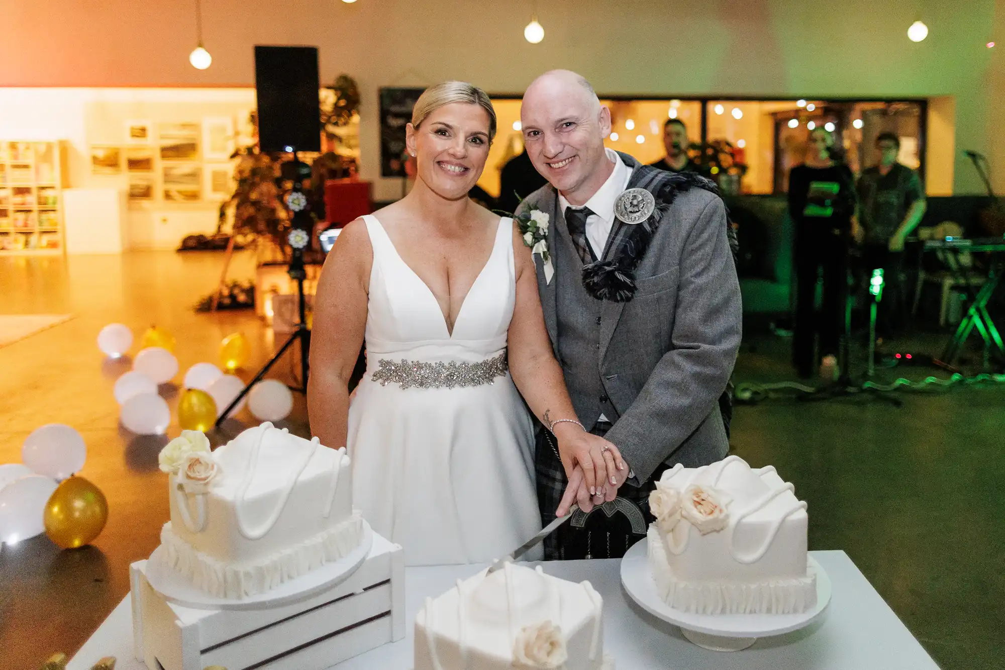 A smiling bride in a white dress and a groom in a grey suit and tartan sash cut a white wedding cake together; three cakes with white icing and roses are on the table. Balloons and fairy lights decorate the modern indoor venue in the background.