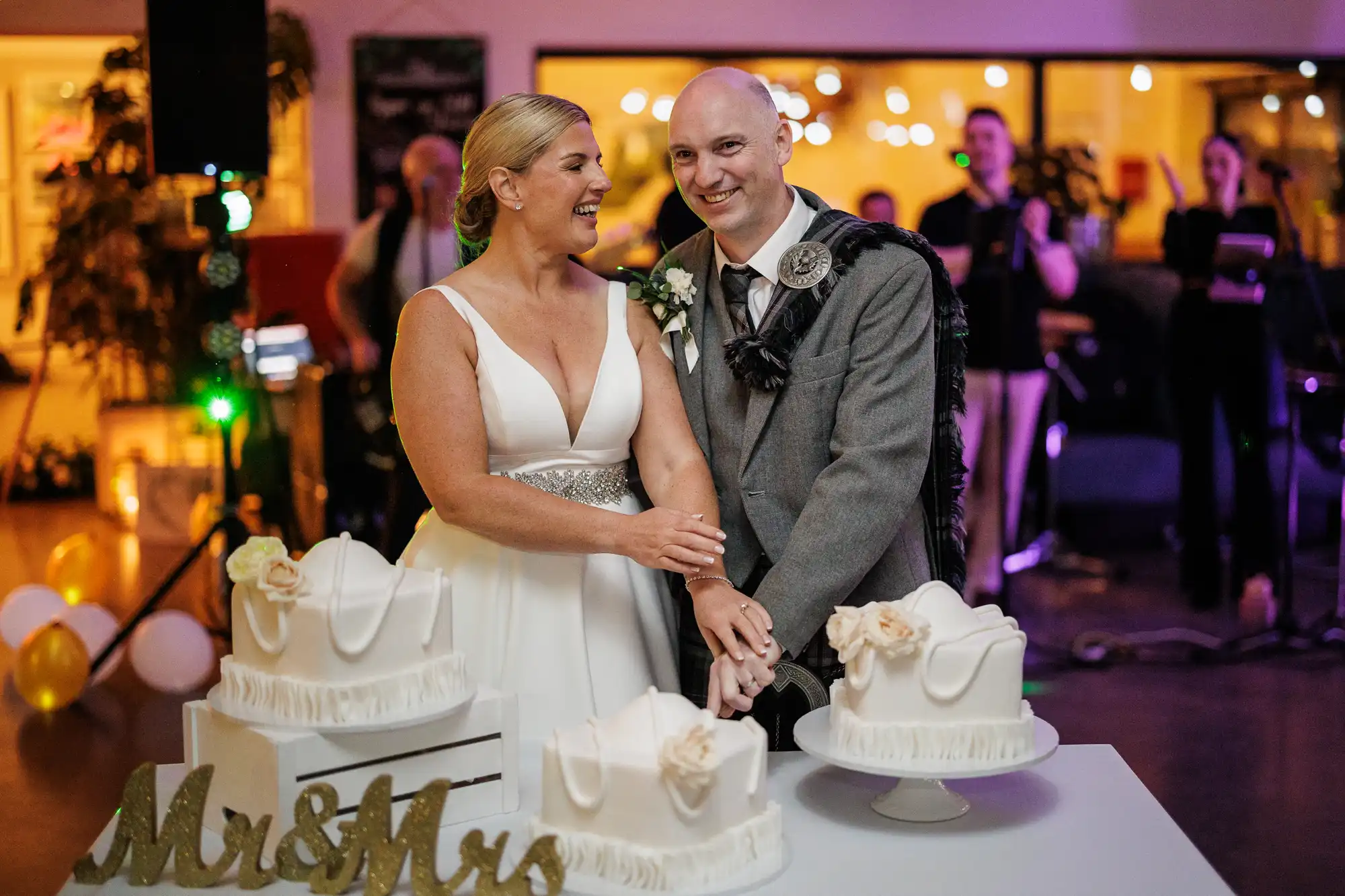 A smiling bride in a white dress and a groom in a grey suit with a tartan sash cut a white wedding cake together. There are two more identical cakes and a gold “Mr & Mrs” sign on the table. People and lights are blurred in the festive background.