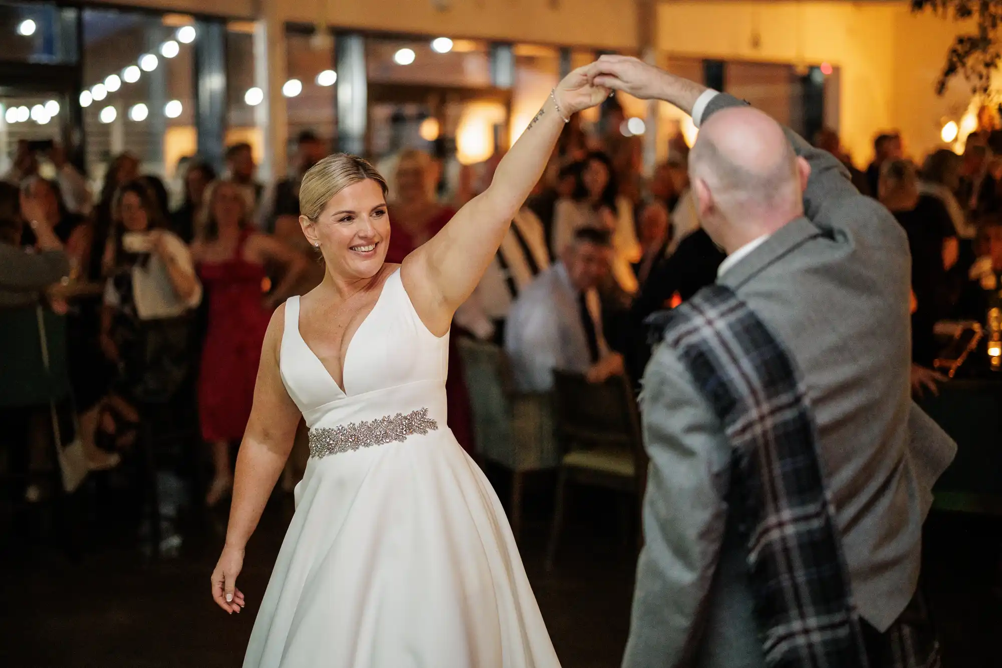 A bride in a white gown with a jewelled belt smiles as she dances, holding hands with a man in a grey suit and checked sash. They are surrounded by seated and standing wedding guests in a warmly lit indoor venue.