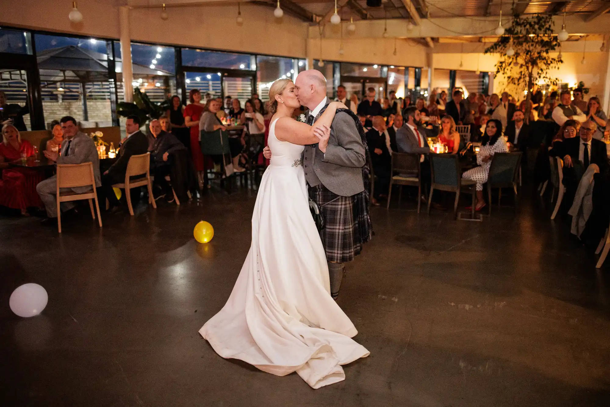A bride in a white dress and a groom in a kilt dance closely in the centre of a warmly lit reception hall, surrounded by seated guests watching them. Balloons lie on the floor, and tables are decorated with candles and glasses.