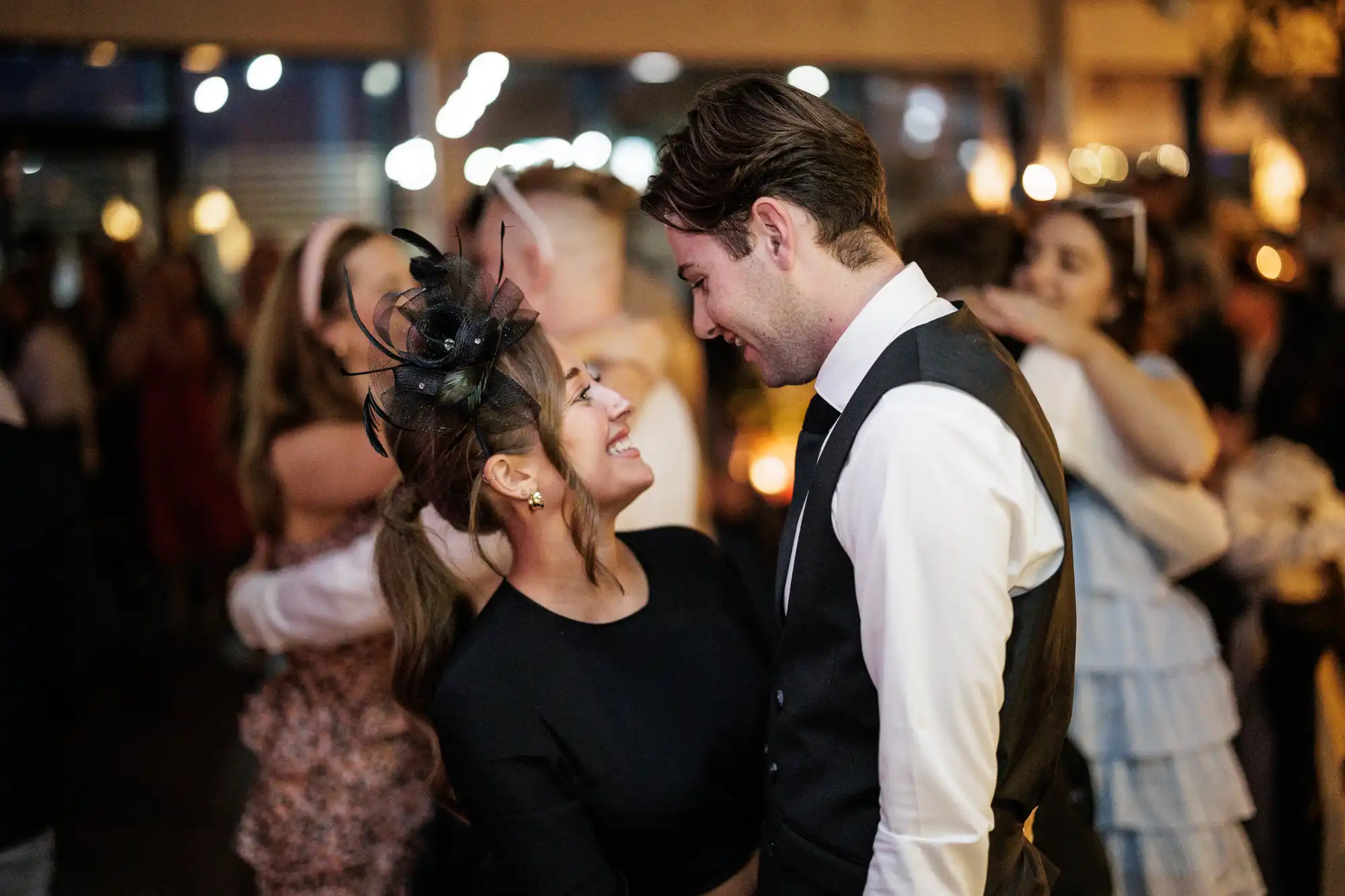 A smiling couple dressed formally dance closely at a lively indoor event. The woman wears a black dress and a decorative fascinator; the man wears a white shirt and dark waistcoat. Blurred guests in festive attire dance and embrace in the warmly lit background.