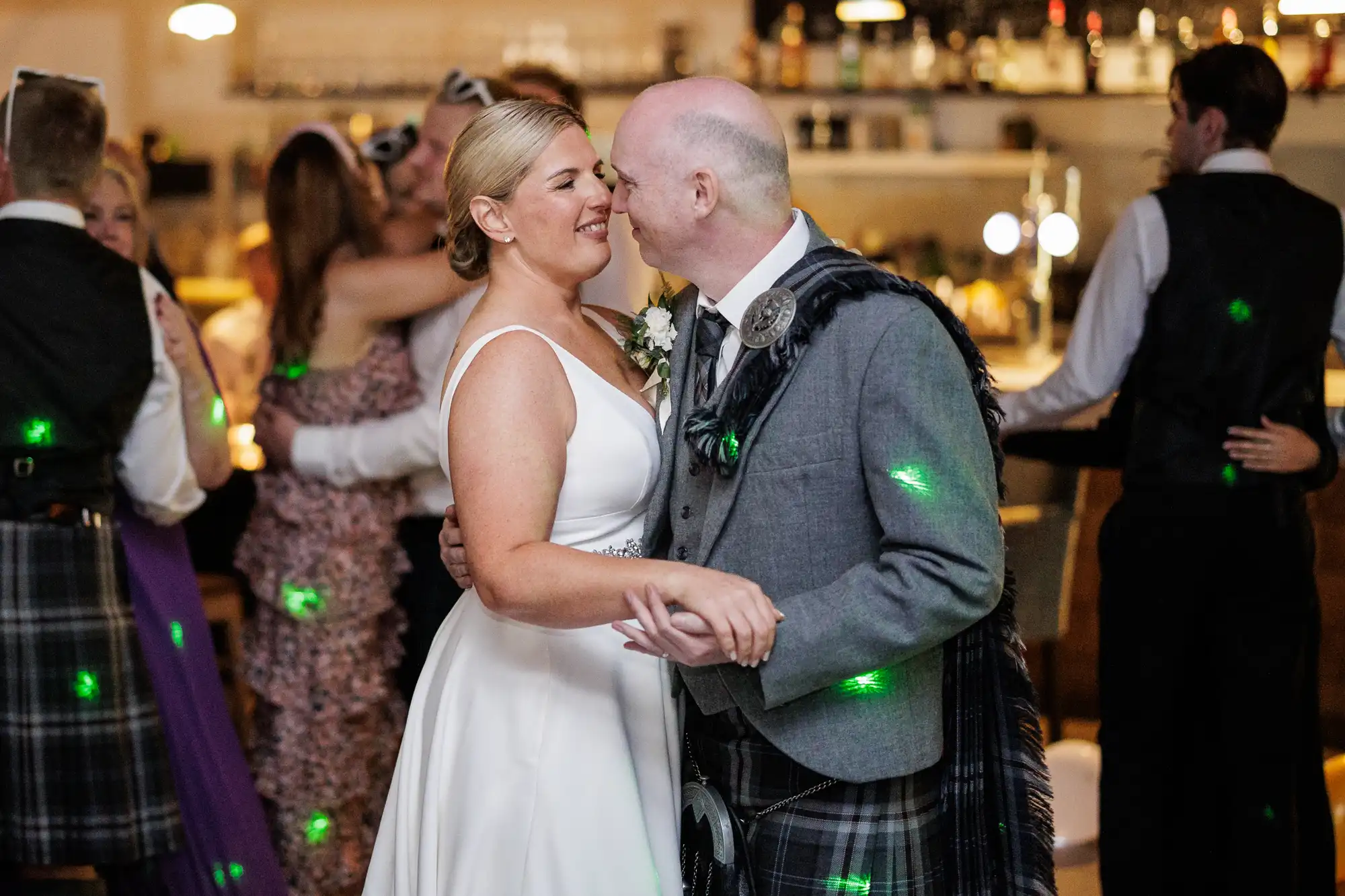 A bride in a white sleeveless gown and a groom in a grey jacket with a kilt smile and dance closely at their wedding reception. Other couples dance in the warmly lit background, and green disco lights dot the scene.